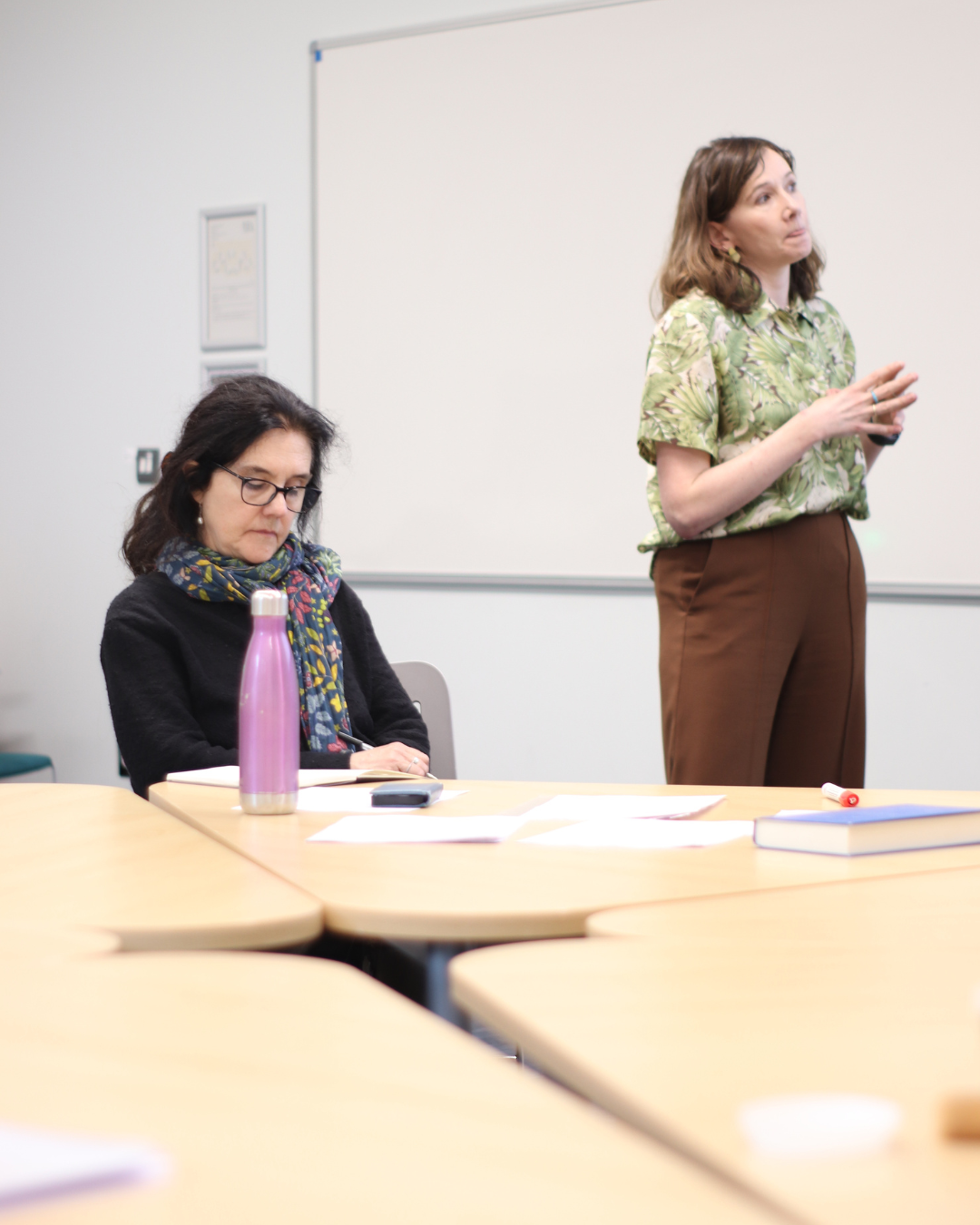 A woman in a green patterned shirt presents in a classroom, while another woman with glasses and a colorful scarf sits at a desk and listens.