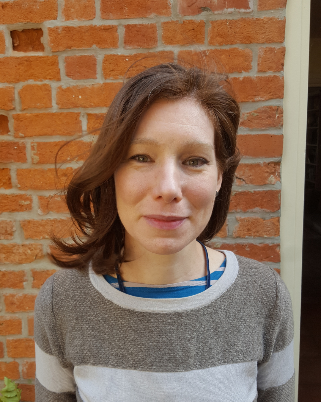 A woman with brown hair and light eyes smiling slightly in front of a brick wall. She wears a grey and white striped sweater over a blue shirt.