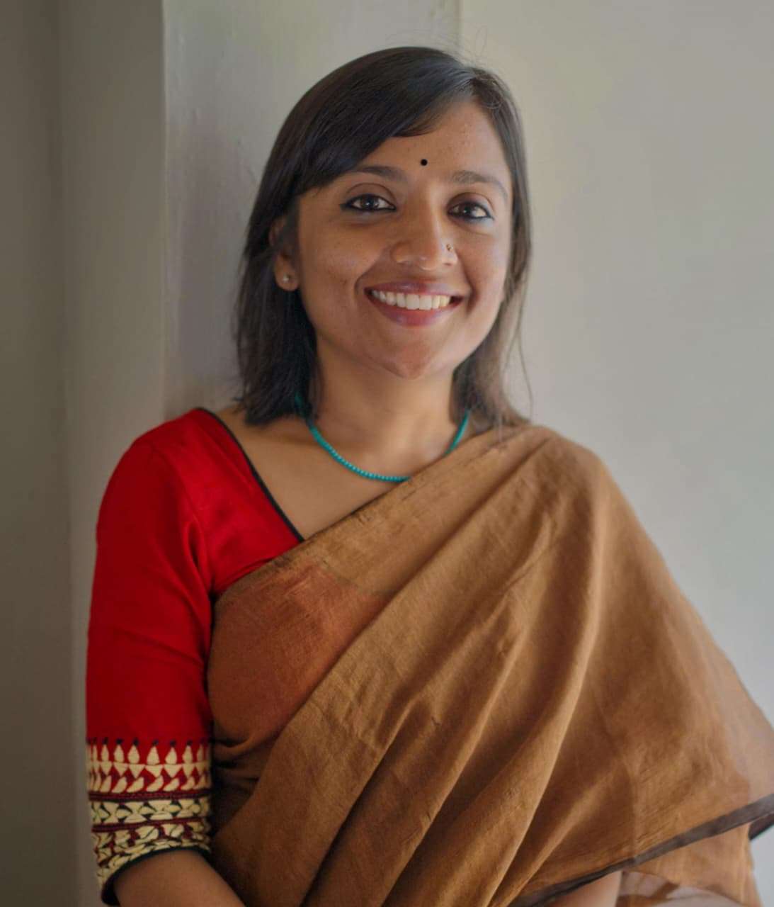 Smiling woman in a brown saree with a red blouse, standing near a light-colored wall.