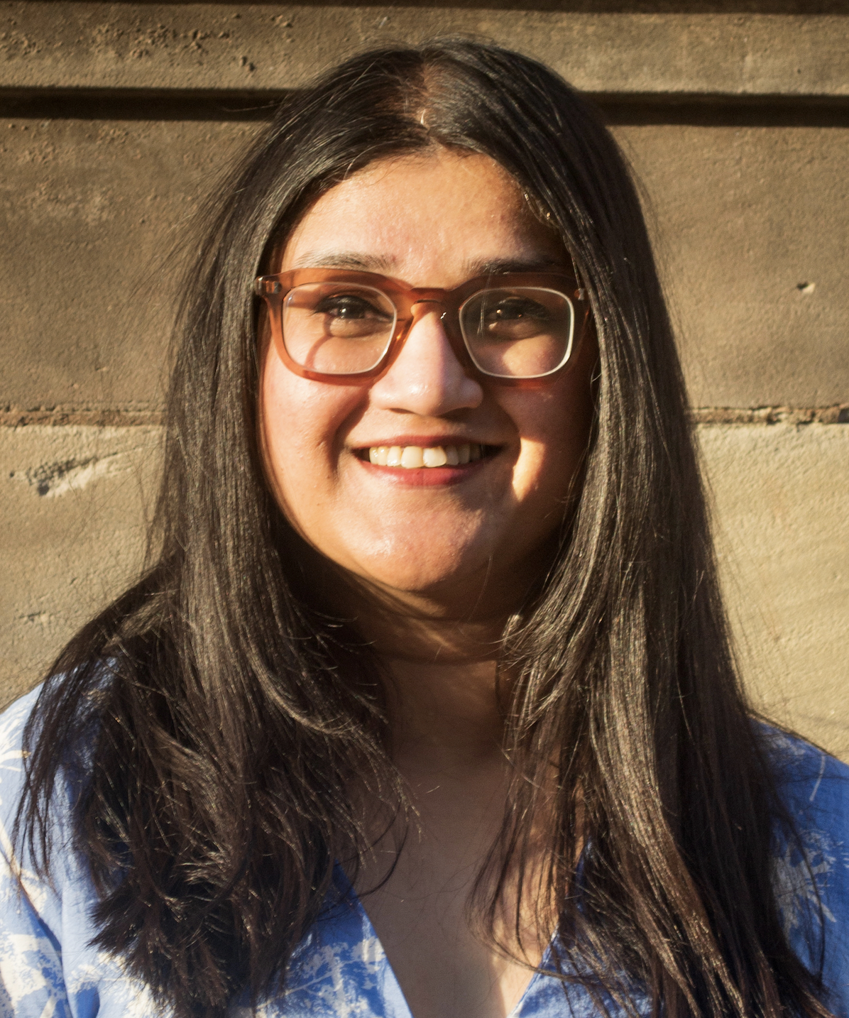 A headshot of a smiling woman with long dark hair and brown-rimmed glasses, standing in front of a textured wall in warm light.