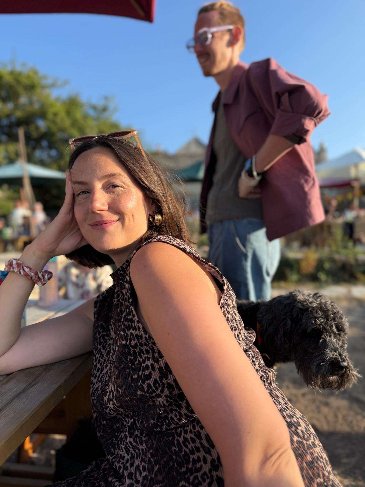Woman smiling at a picnic table, wearing a leopard print dress. A small black dog is beside her, and a man stands in the background.