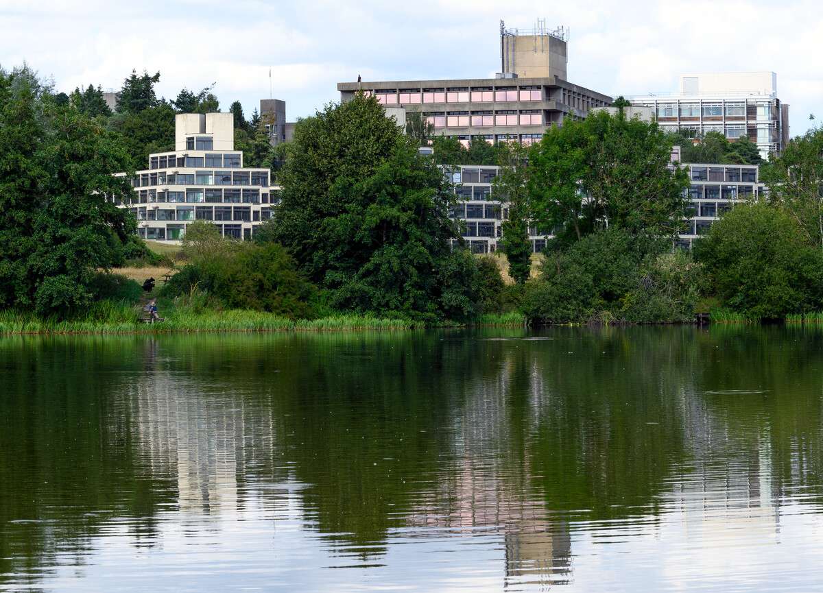Modern university buildings nestled among green trees on a hillside, with their reflections mirrored in the calm lake in the foreground.
