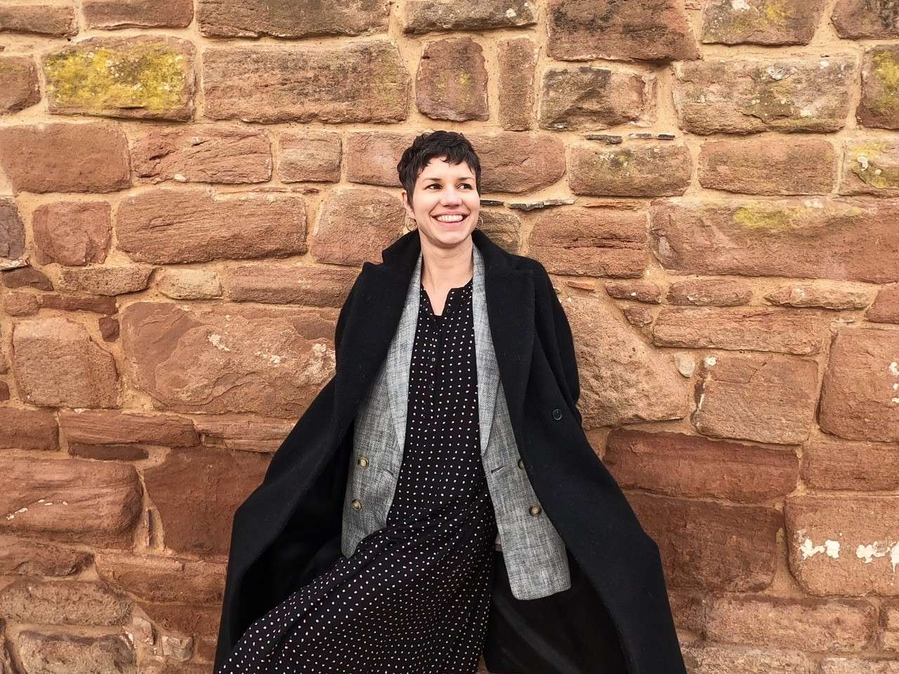 A person smiling, wearing a black coat and polka dot dress, leans casually against a stone wall.