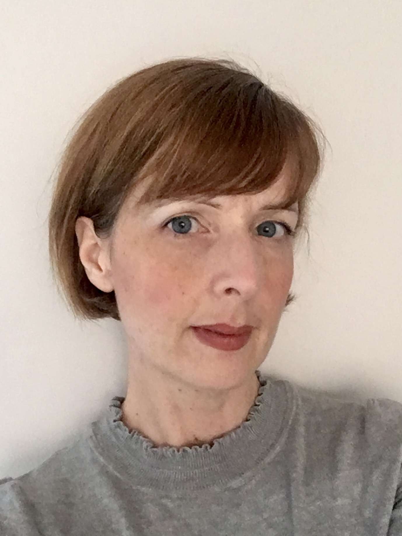A headshot of a woman with short, reddish-brown hair and blue eyes. She wears a grey sweater with a ruffled collar against a plain background.