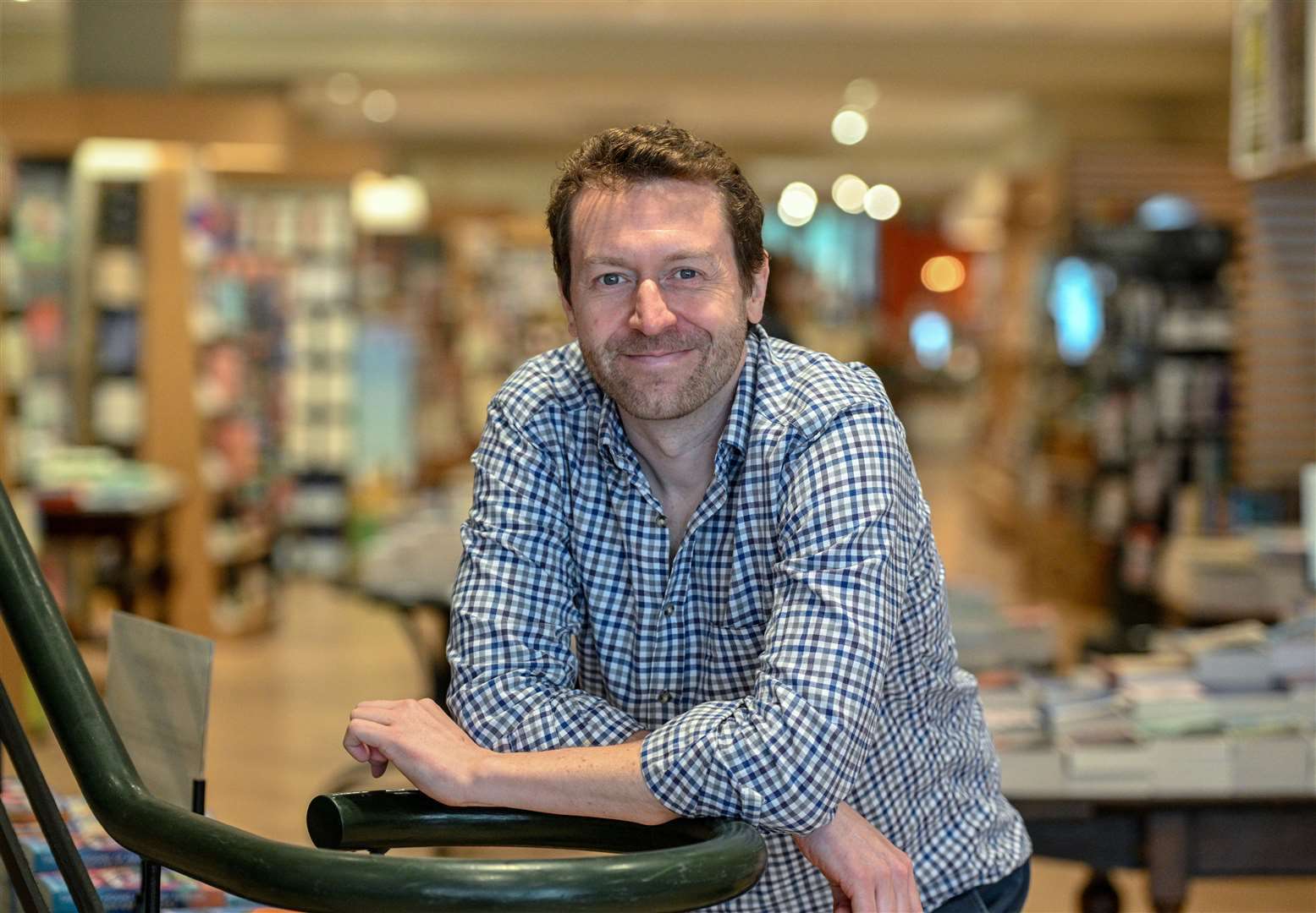 A man in a blue checkered shirt smiles as he leans on a chair in a bookstore, with bookshelves blurred in the background.