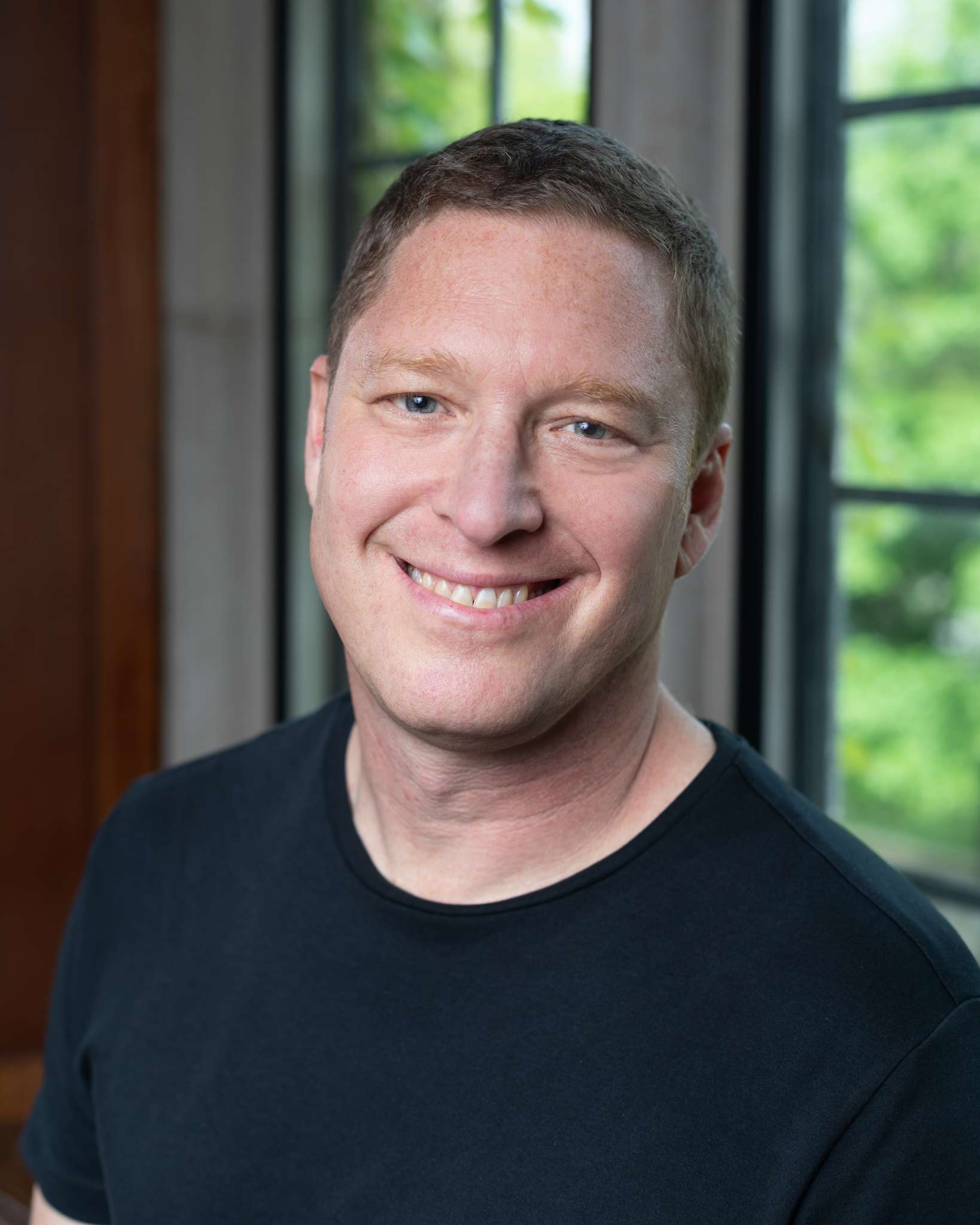A headshot of a smiling man with short brown hair and blue eyes. He is wearing a black t-shirt and is indoors by a window with greenery outside.