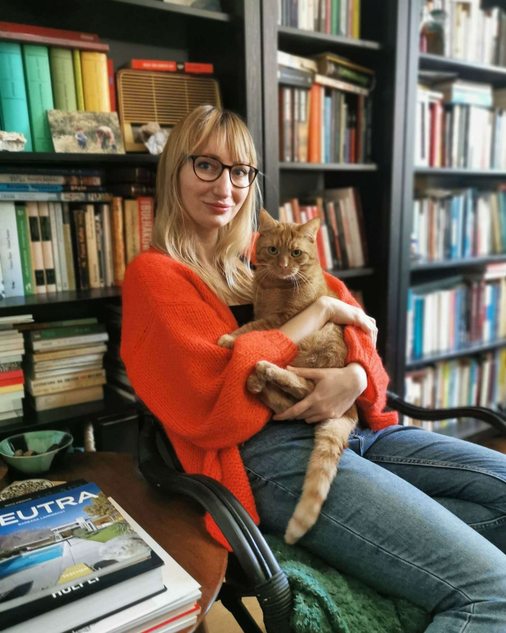 Person in glasses and red sweater holding an orange cat, sitting in a chair, surrounded by bookshelves filled with books.