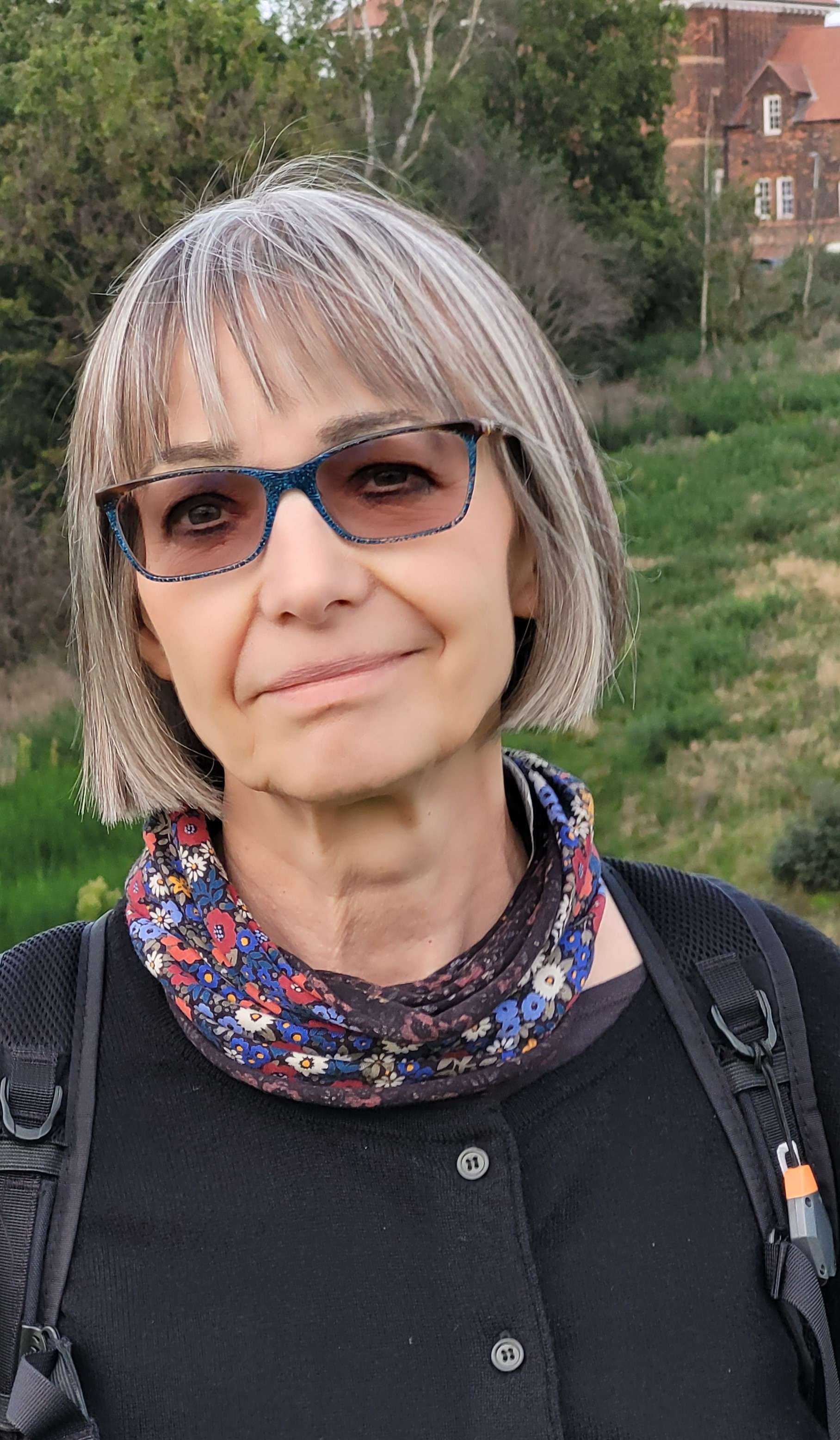 A woman with a short silver bob and blue glasses smiles gently outdoors. She wears a floral scarf and a black backpack.