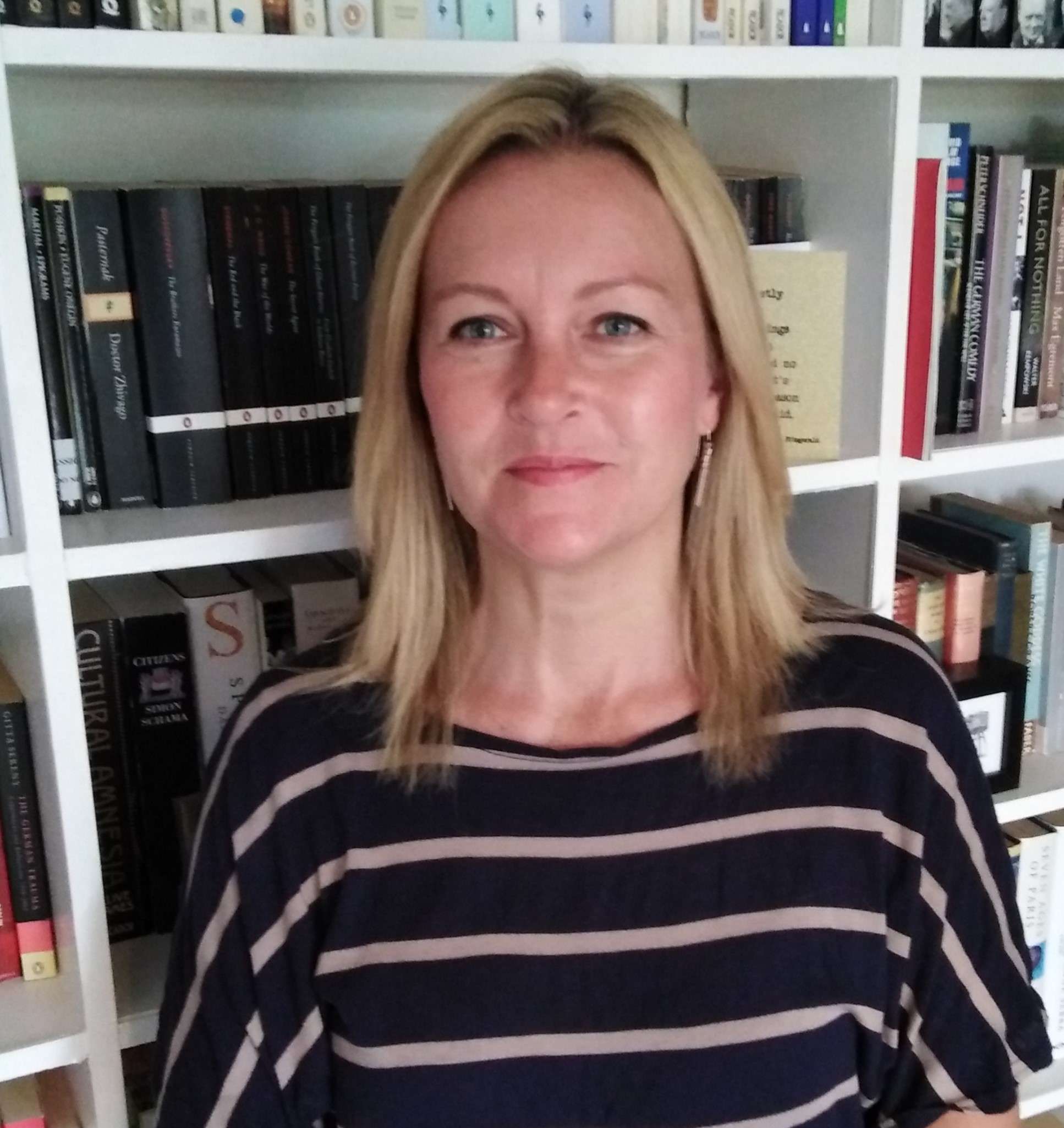 A blonde woman in a striped shirt looks at the camera, standing in front of a white bookshelf.