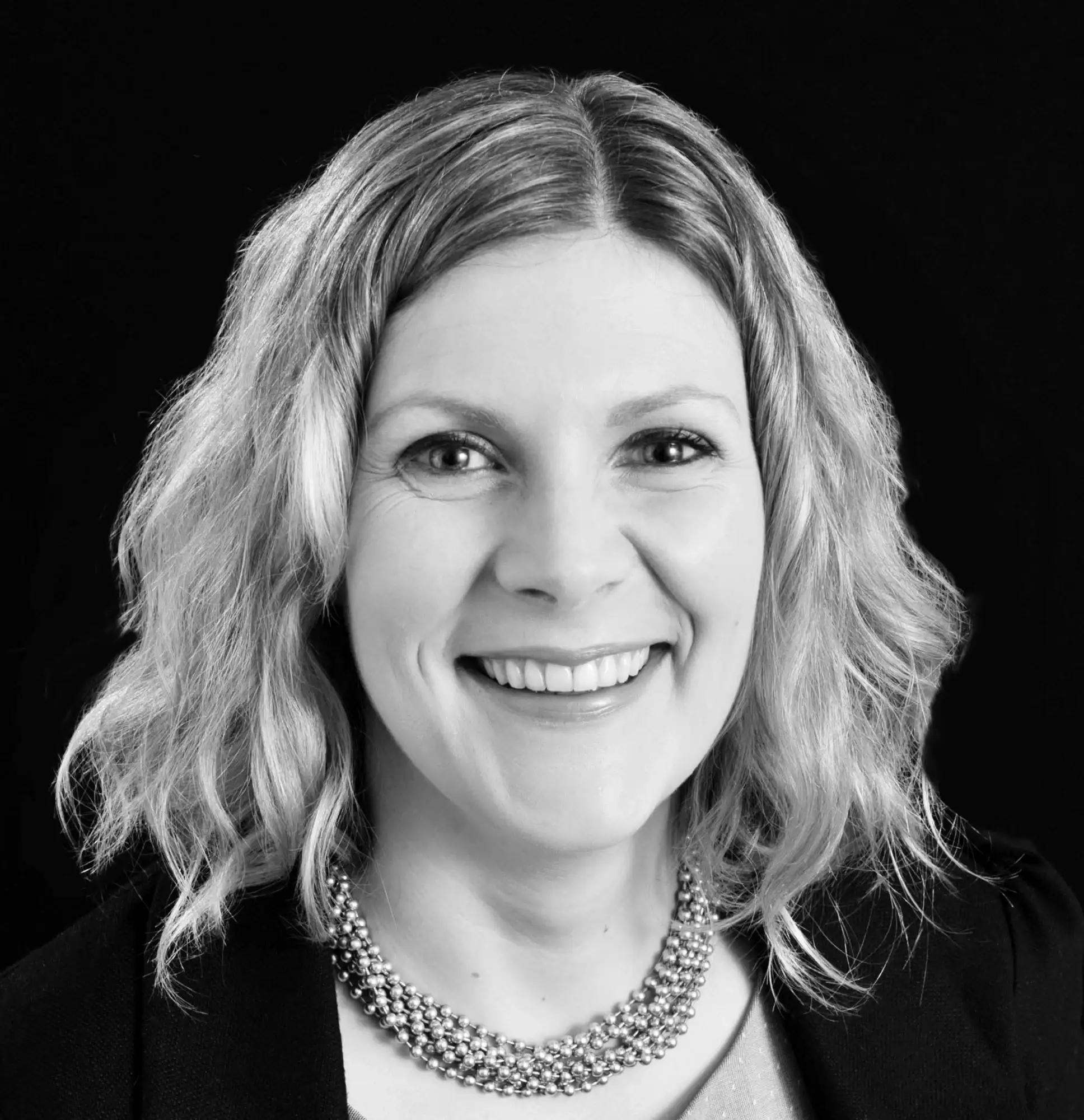 A black and white headshot of a smiling woman with wavy, shoulder-length hair and a multi-strand beaded necklace, set against a black background.