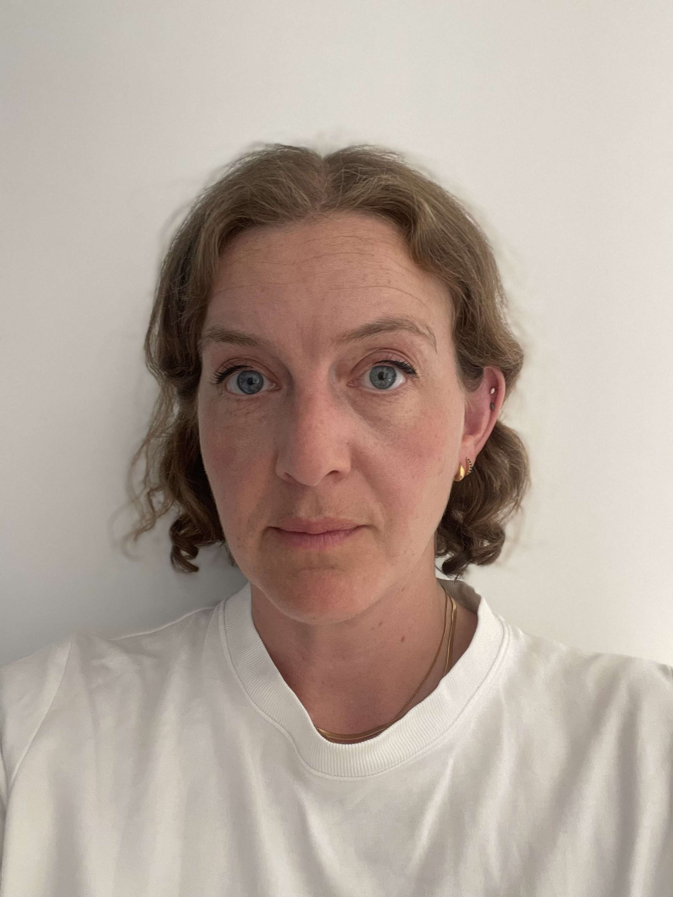 A person with short, wavy brown hair and blue eyes, wearing a white shirt, looks directly at the camera against a plain backdrop.