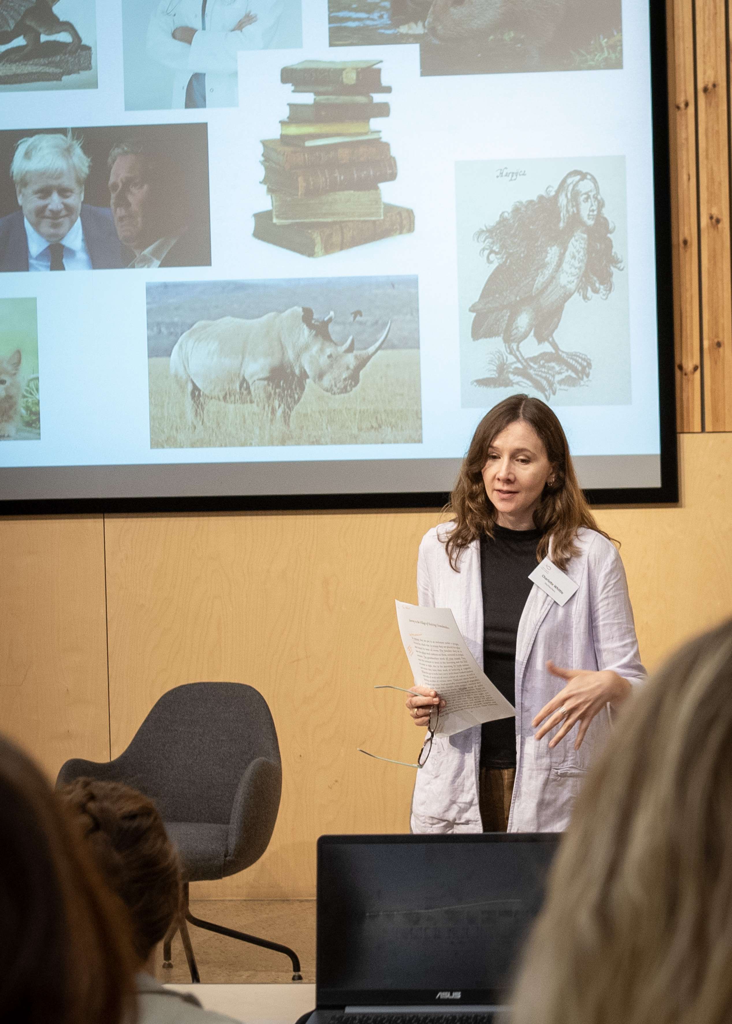 A woman gives a presentation, standing before a screen with a collage of images including Boris Johnson, a rhino, old books, and a drawing of a harpy.