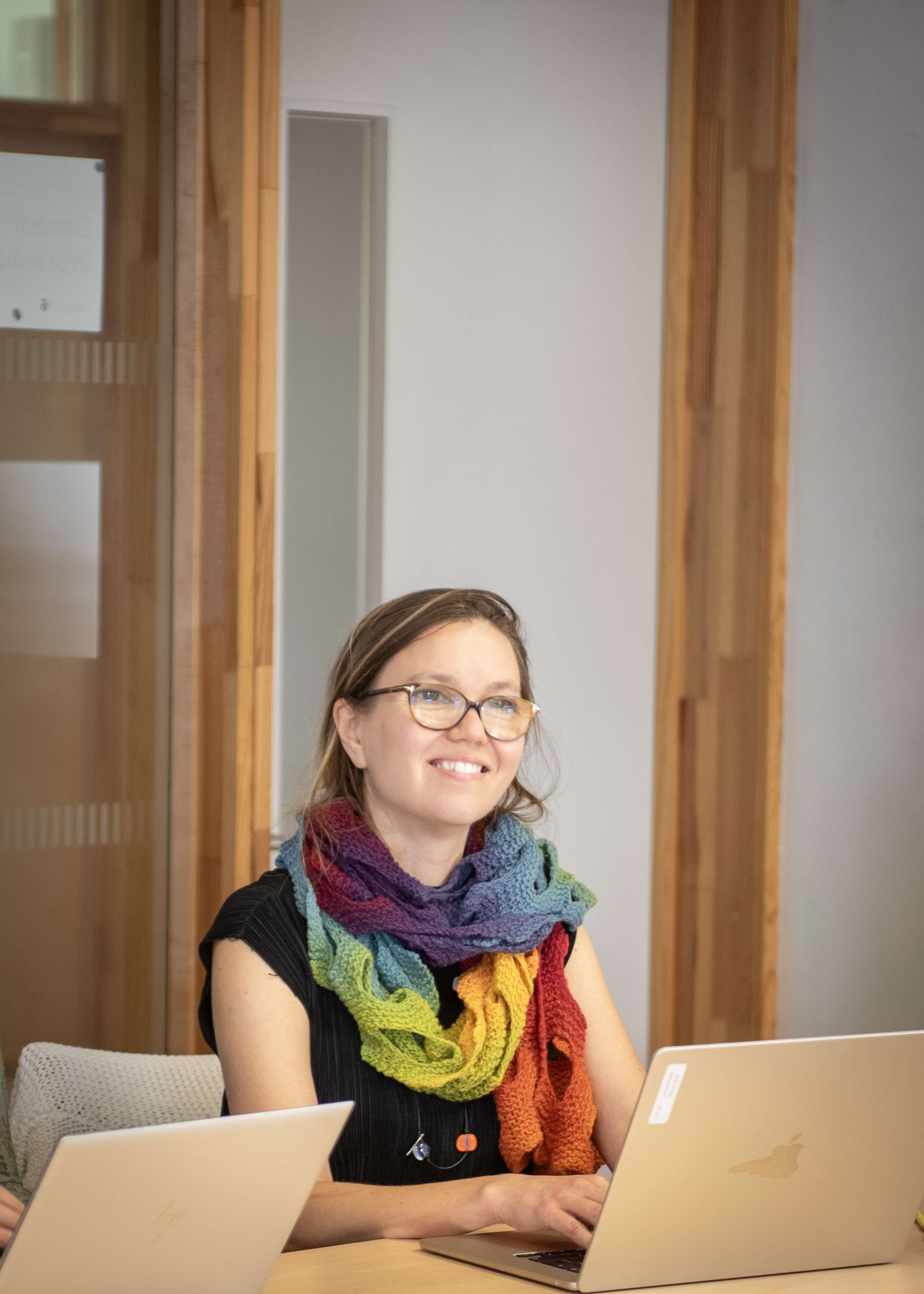 A smiling woman wearing glasses and a bright, rainbow-colored knitted scarf sits at a table with laptops, looking up and to the side.