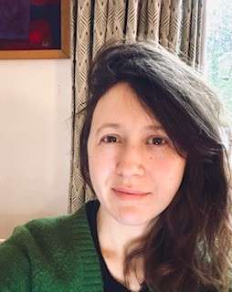 A headshot of a woman with long, dark wavy hair and a slight smile, wearing a green knit cardigan in front of a window.