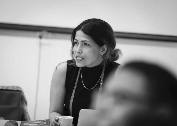 Black and white photo of a woman seated indoors, holding a cup, looking intently at someone, with a blurred foreground figure.