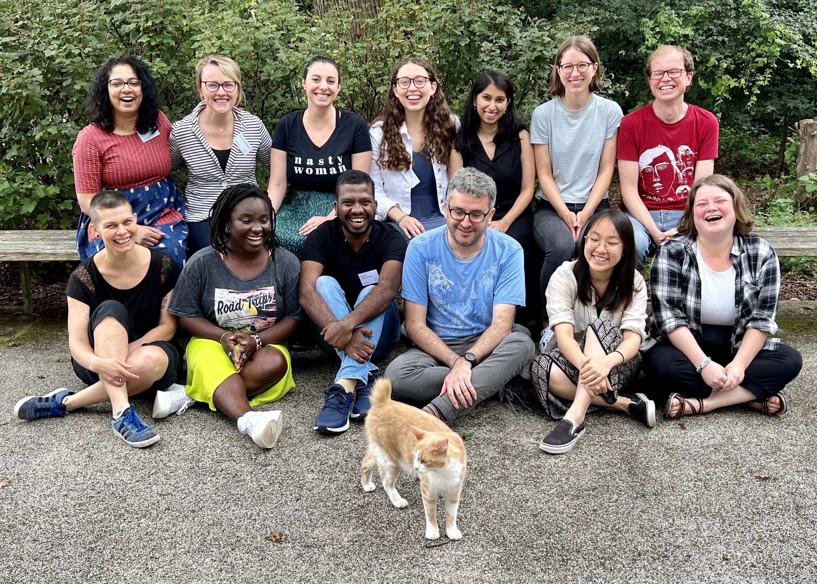 A diverse group of thirteen smiling people pose for a photo outdoors in front of some bushes, with a ginger and white cat standing in the foreground.