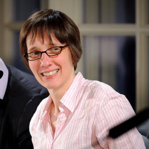 A person with short brown hair and glasses, wearing a striped shirt, smiles while seated indoors, with a blurred background.