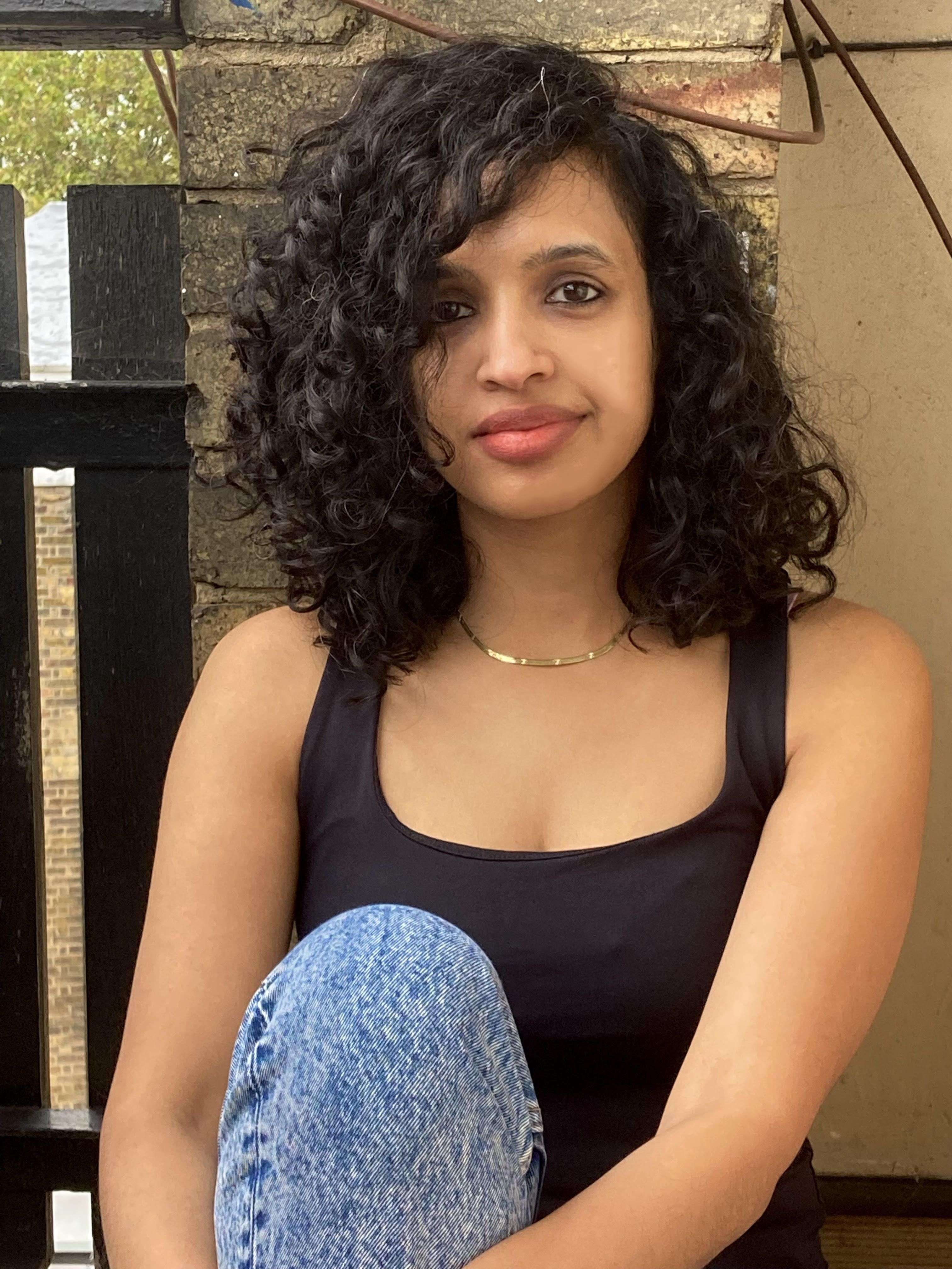 A young woman with dark curly hair looks at the camera. She wears a black tank top, blue jeans, and a gold necklace, sitting by a dark fence.