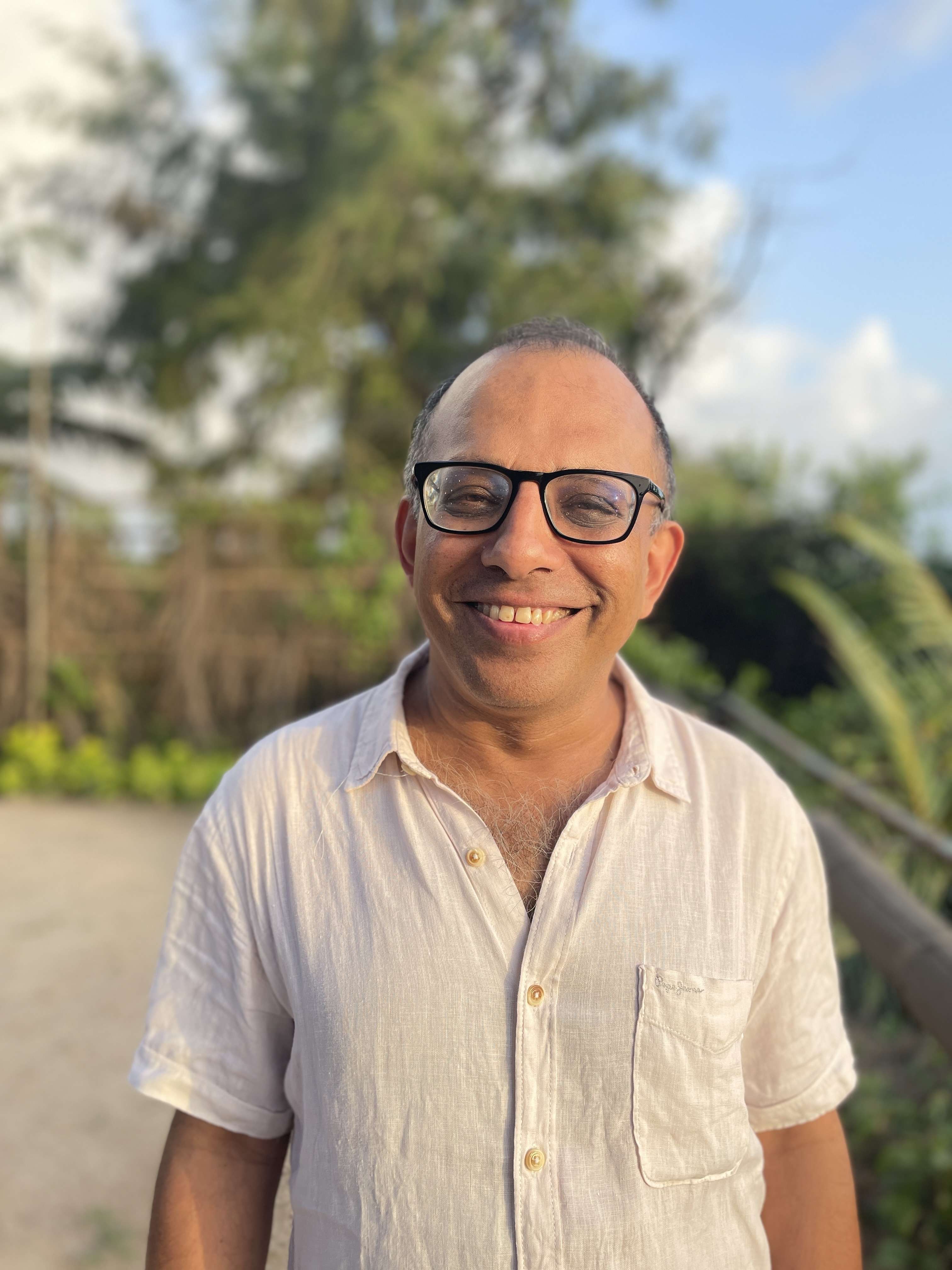 A man with glasses and a light-colored, button-down shirt smiles warmly at the camera while standing outdoors.
