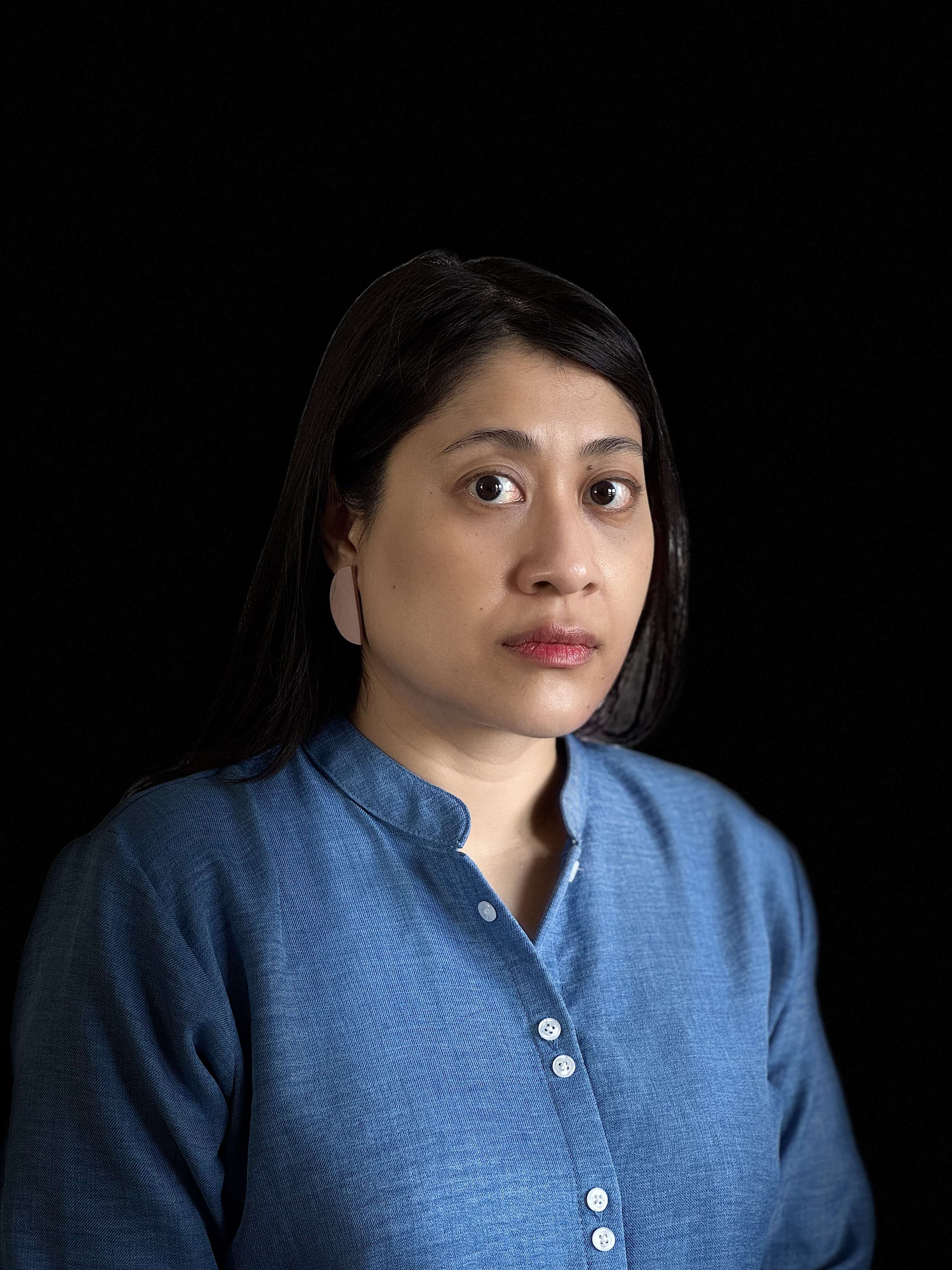 An Indonesian woman with long, black hair and pink, half-moon earrings, wearing a blue, button-down shirt with a round collar, looking to the camera.
