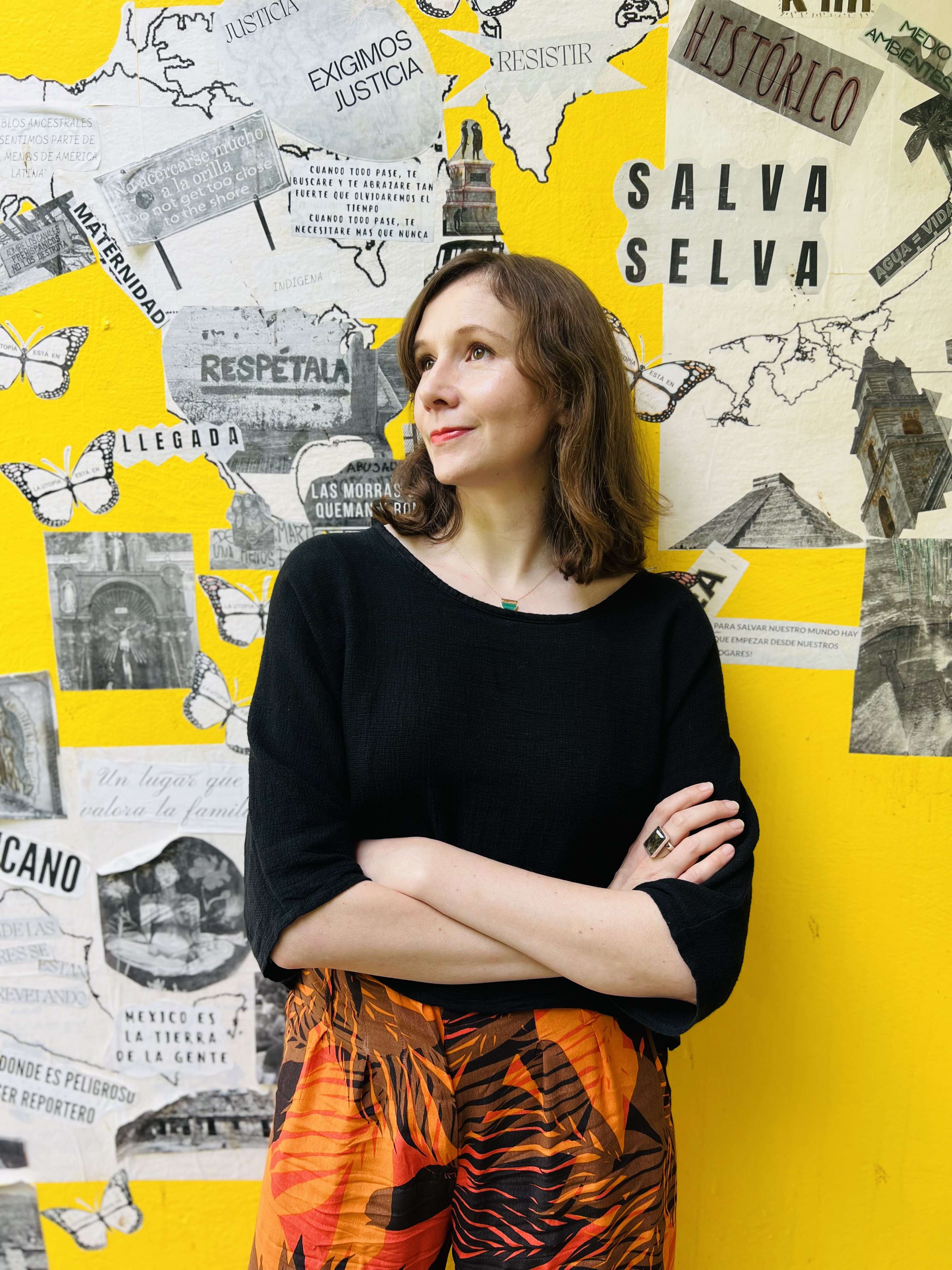 A person in a black shirt and vibrant skirt stands against a yellow wall with various collage elements including maps, text, and butterflies.