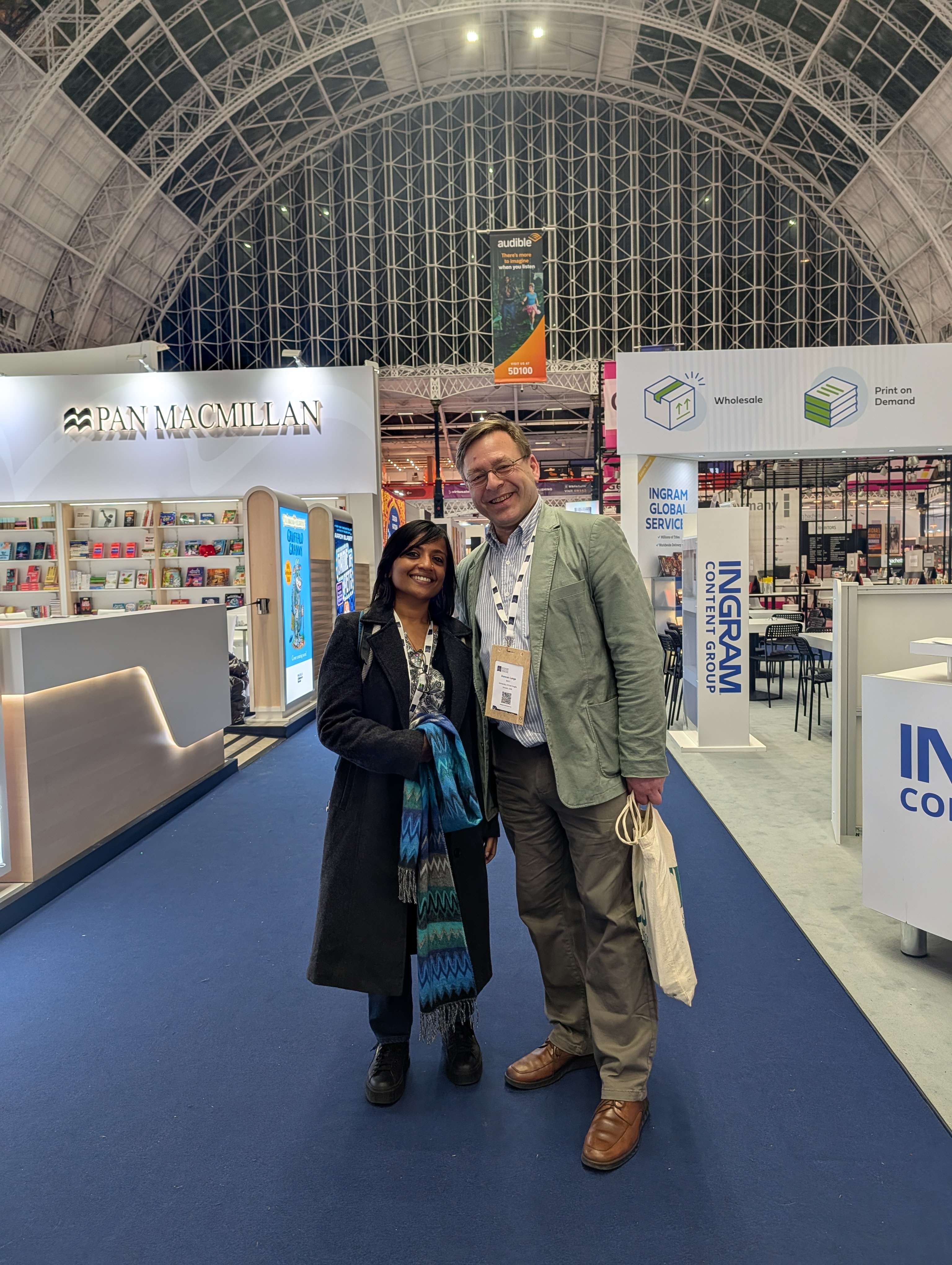 A man and a woman smile for a photo in a large convention hall with an arched ceiling, standing between Pan Macmillan and Ingram exhibition booths.