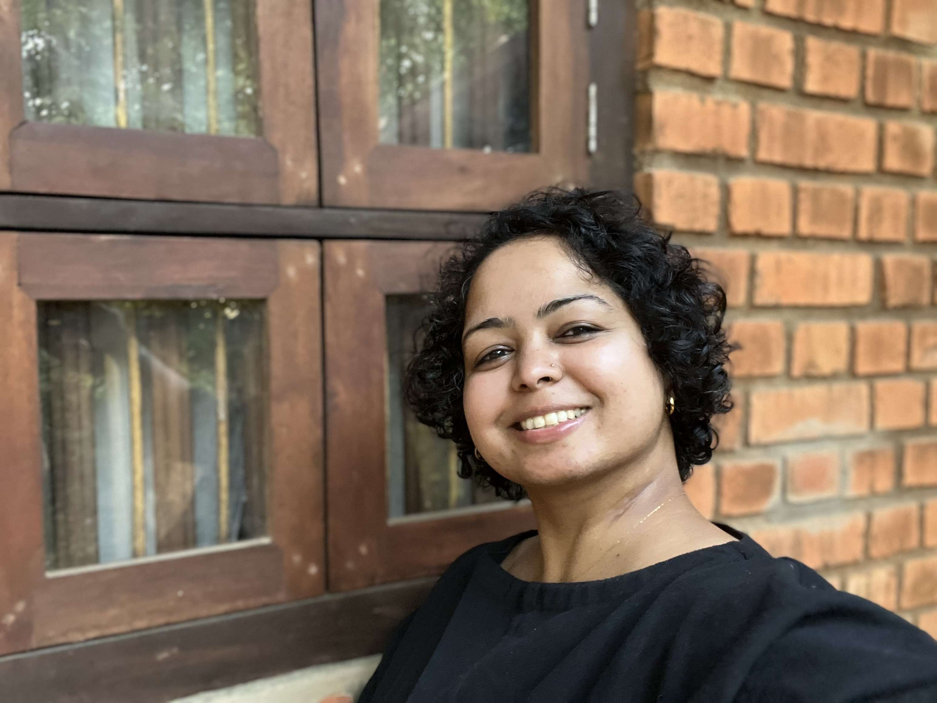 A smiling woman with short, dark curly hair poses in a black top, with a wooden-framed window and a red brick wall in the background.