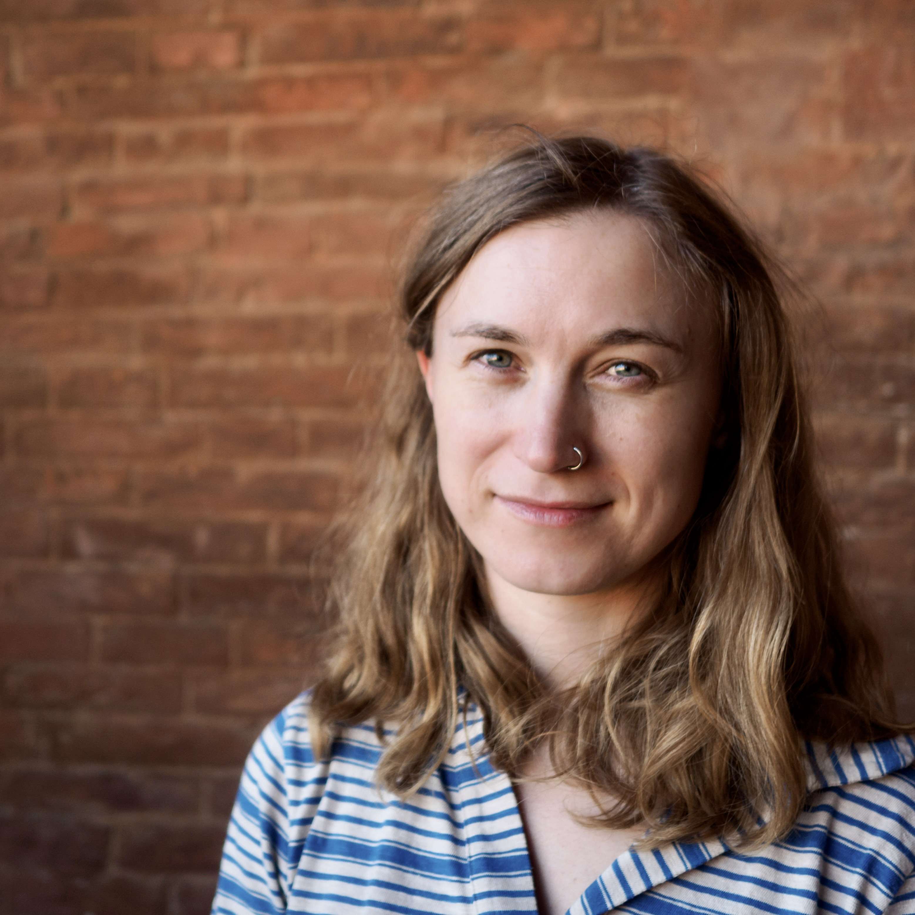 A headshot of a woman with wavy, light-brown hair and a nose ring. She wears a blue and white striped shirt and smiles in front of a brick wall.