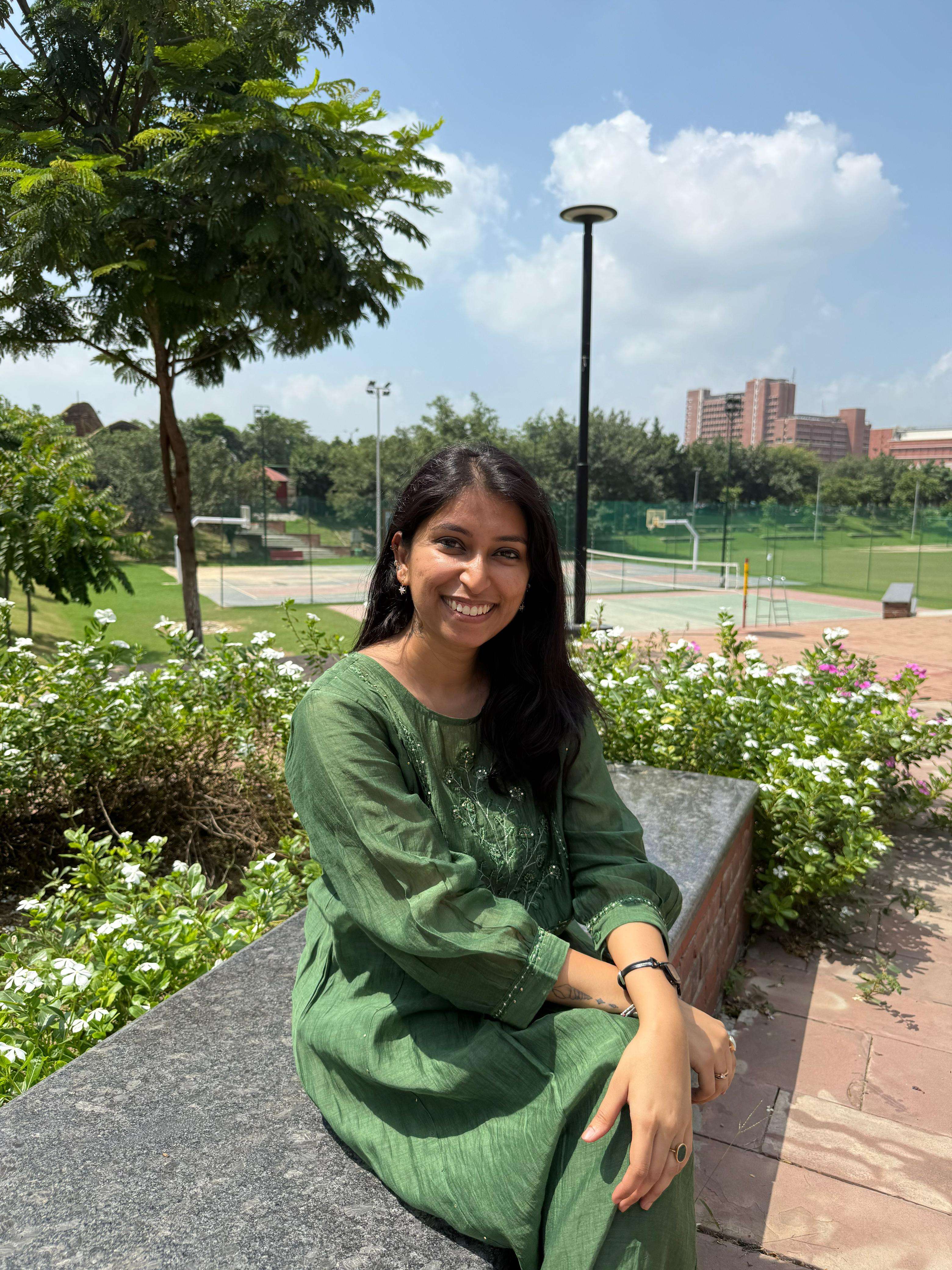 A young woman in a green embroidered tunic smiles while sitting on a stone bench in a park, with tennis courts and trees in the background.