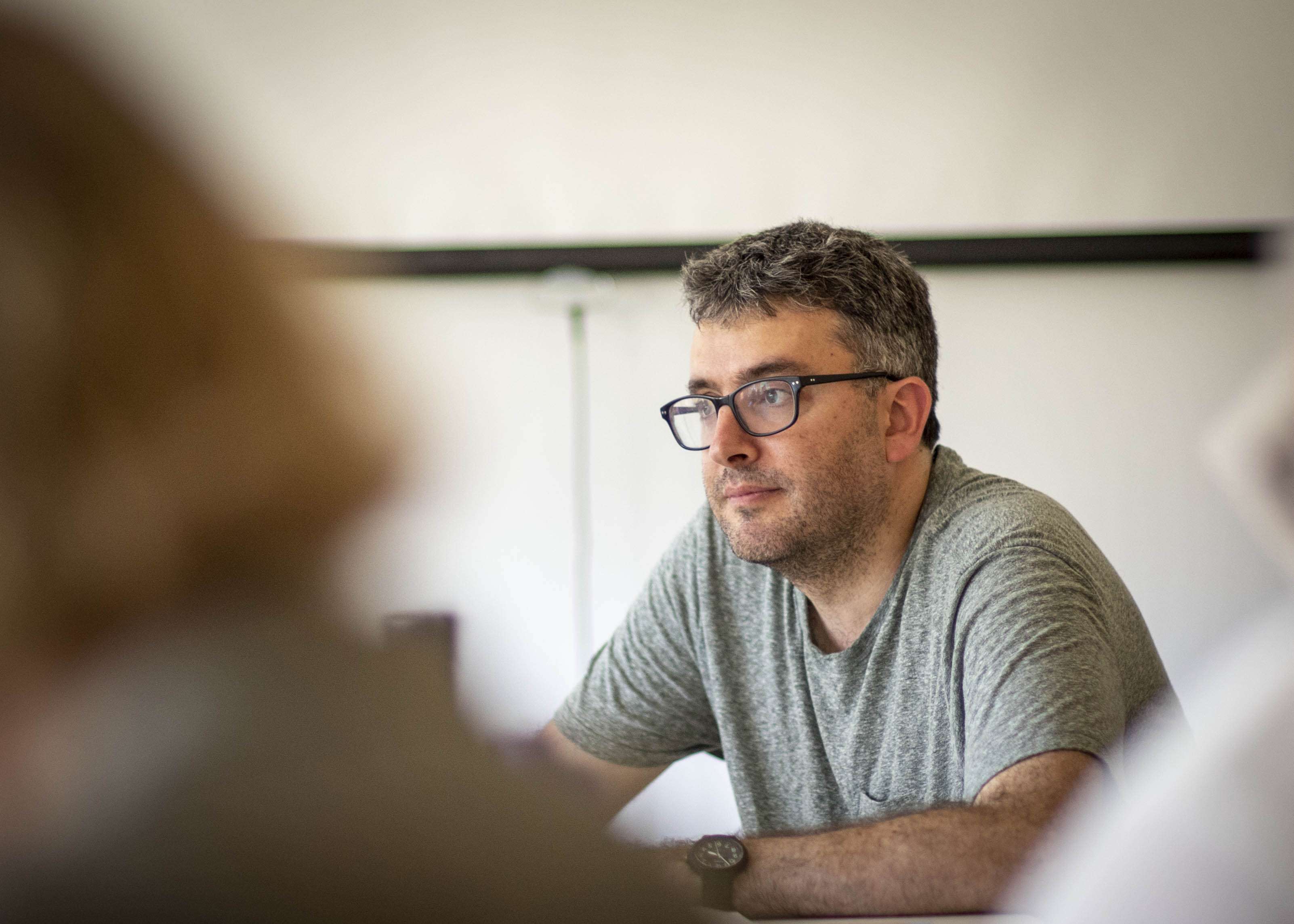A man with glasses and a gray t-shirt sits at a table, looking ahead thoughtfully. The background is blurred.