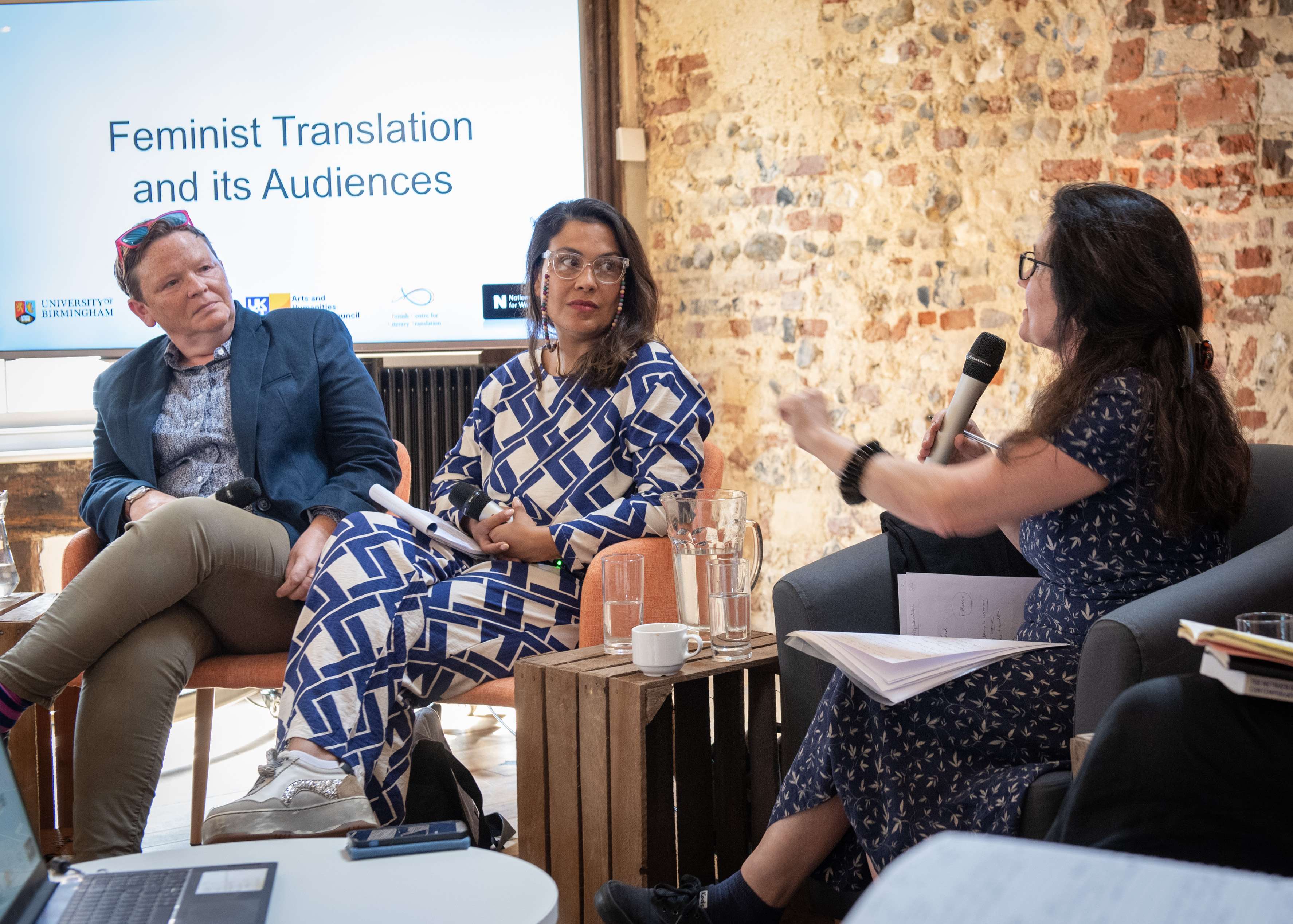 Three people sit on a panel in front of a screen reading "Feminist Translation." One woman speaks into a microphone while the other two listen.