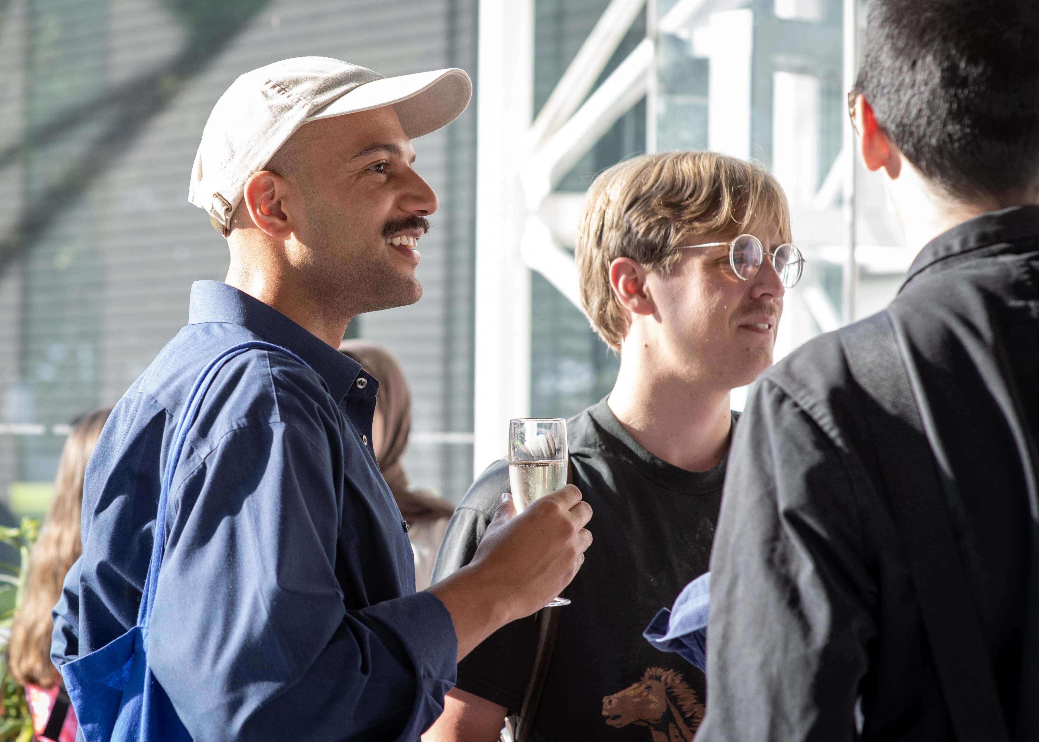 A smiling man in a white cap and blue shirt holds a glass of champagne while talking with two other men at a brightly lit social gathering.
