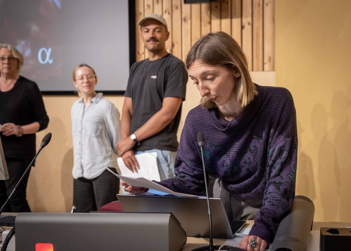 A woman speaks at a podium, reading from papers. Three people stand in the background, one of whom is smiling.