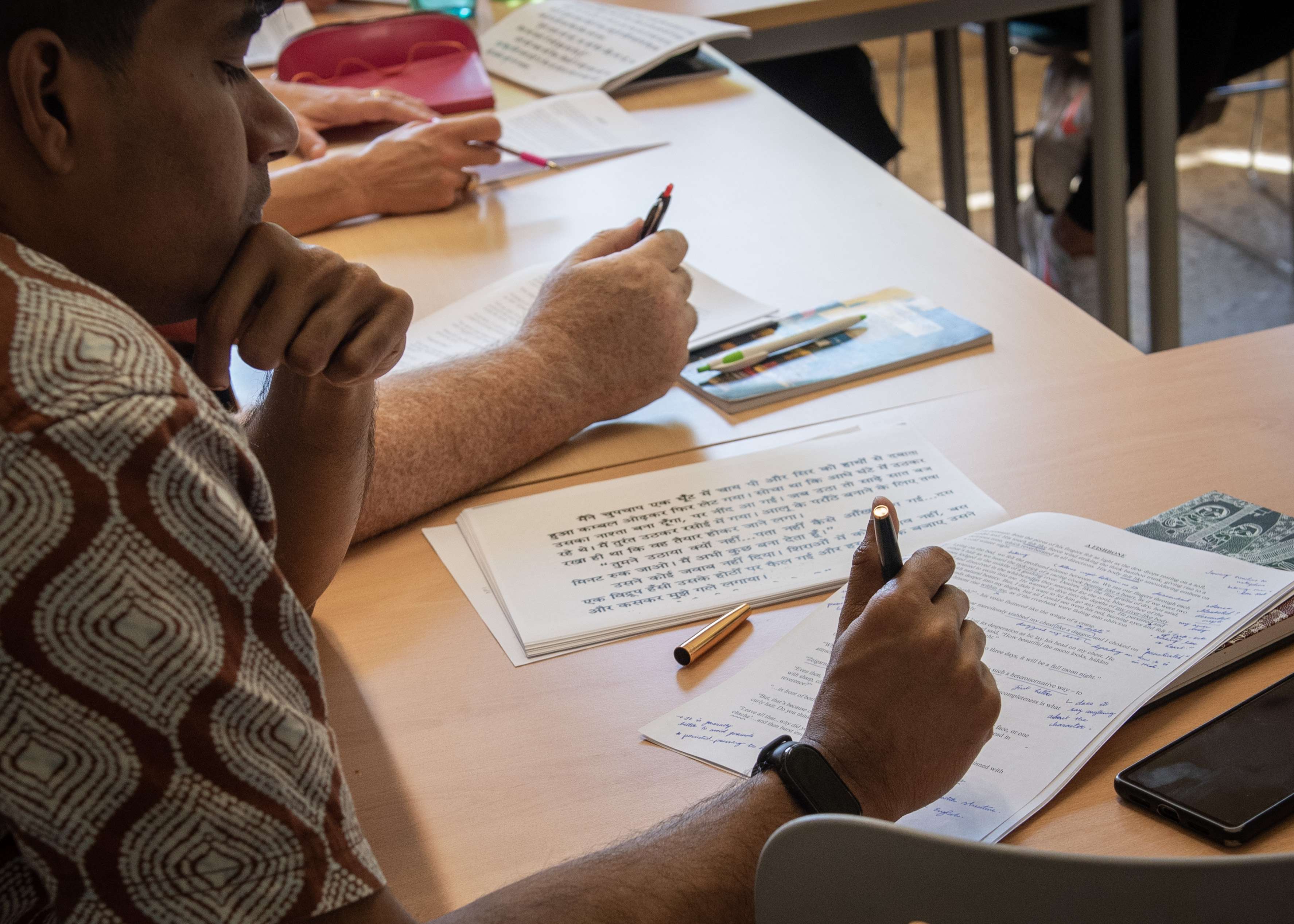 Students in a language class study at a desk, writing notes on papers with text in both English and a non-Latin, possibly Devanagari, script.