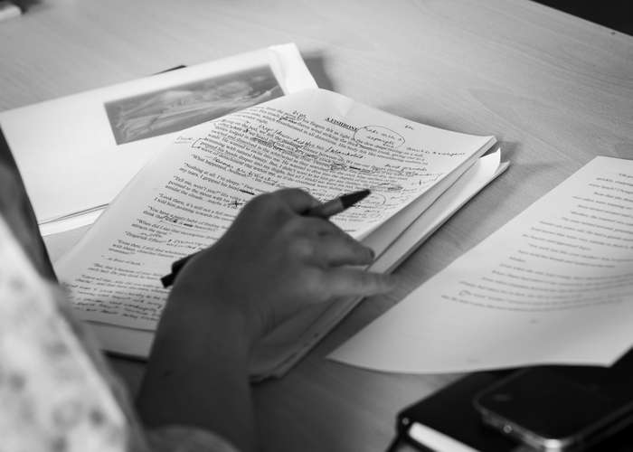 A black and white close-up of a hand holding a pen and marking edits on a typed manuscript resting on a desk.