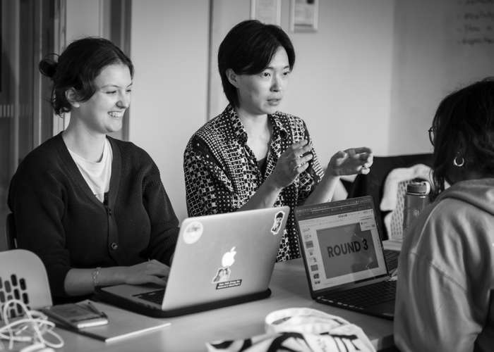 Three people sit at a table with laptops, engaged in discussion. One gestures while speaking. The screen displays "ROUND 3." Black and white image.