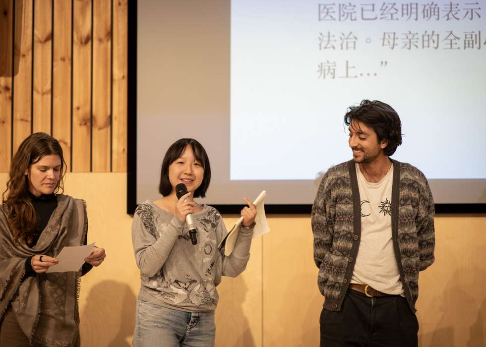 A woman speaks into a microphone at a presentation, standing between another woman reading a paper and a man smiling at her.