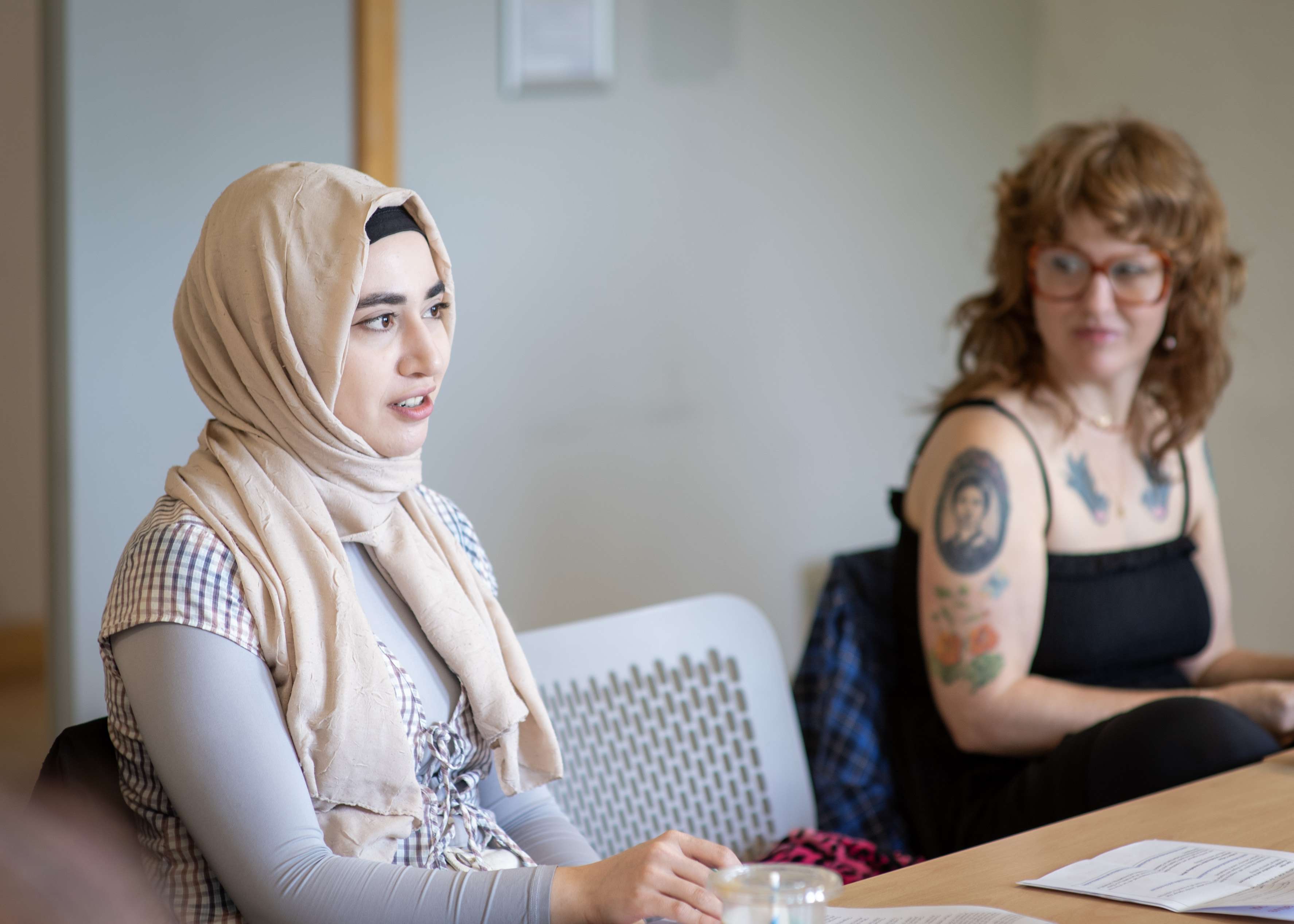 A young woman in a beige hijab speaks while seated at a table. Another woman with glasses and tattoos is blurred in the background.