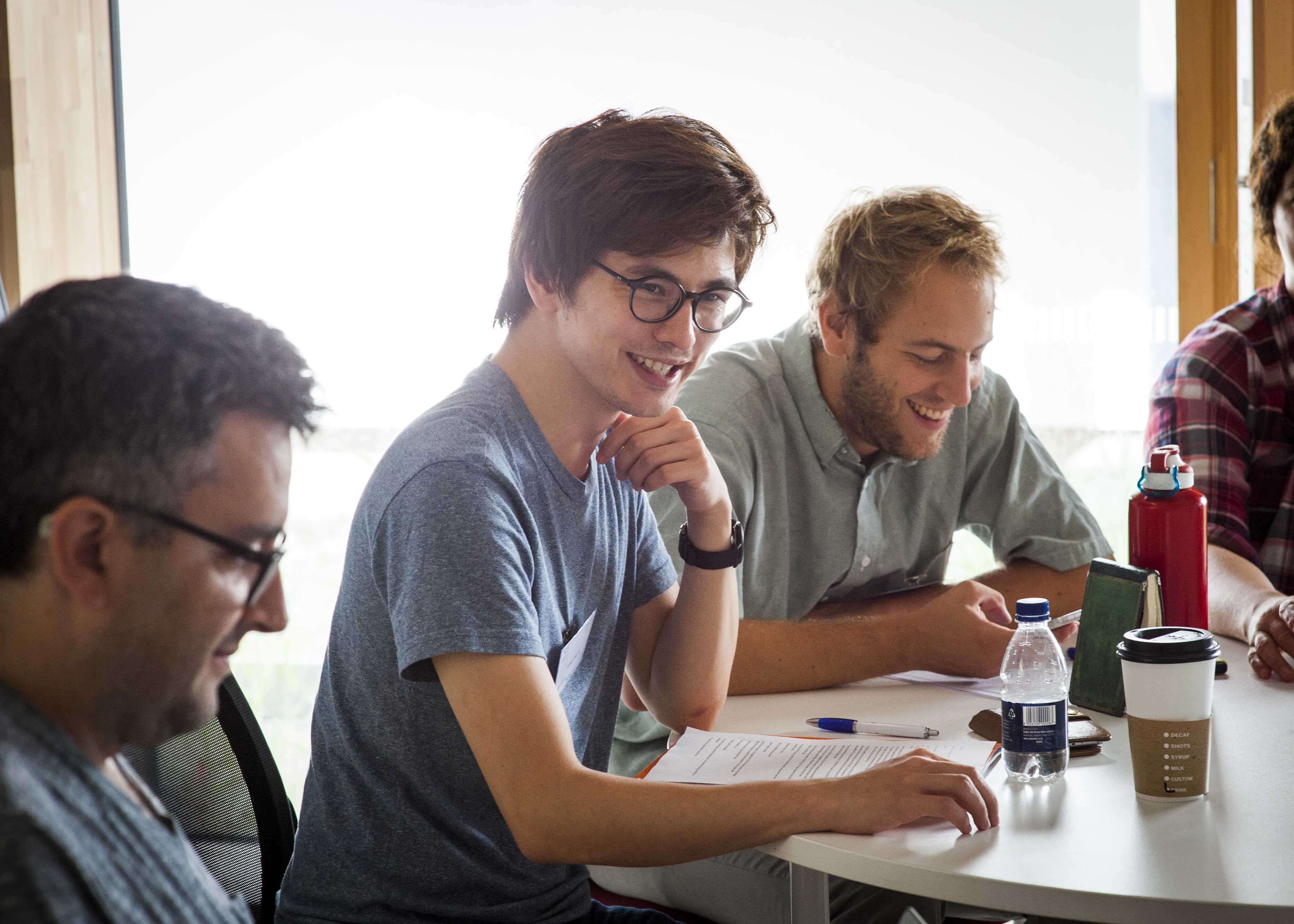 Group of four people sitting around a table, engaged in discussion, with papers, pens, and drinks. One person is smiling and wearing glasses.