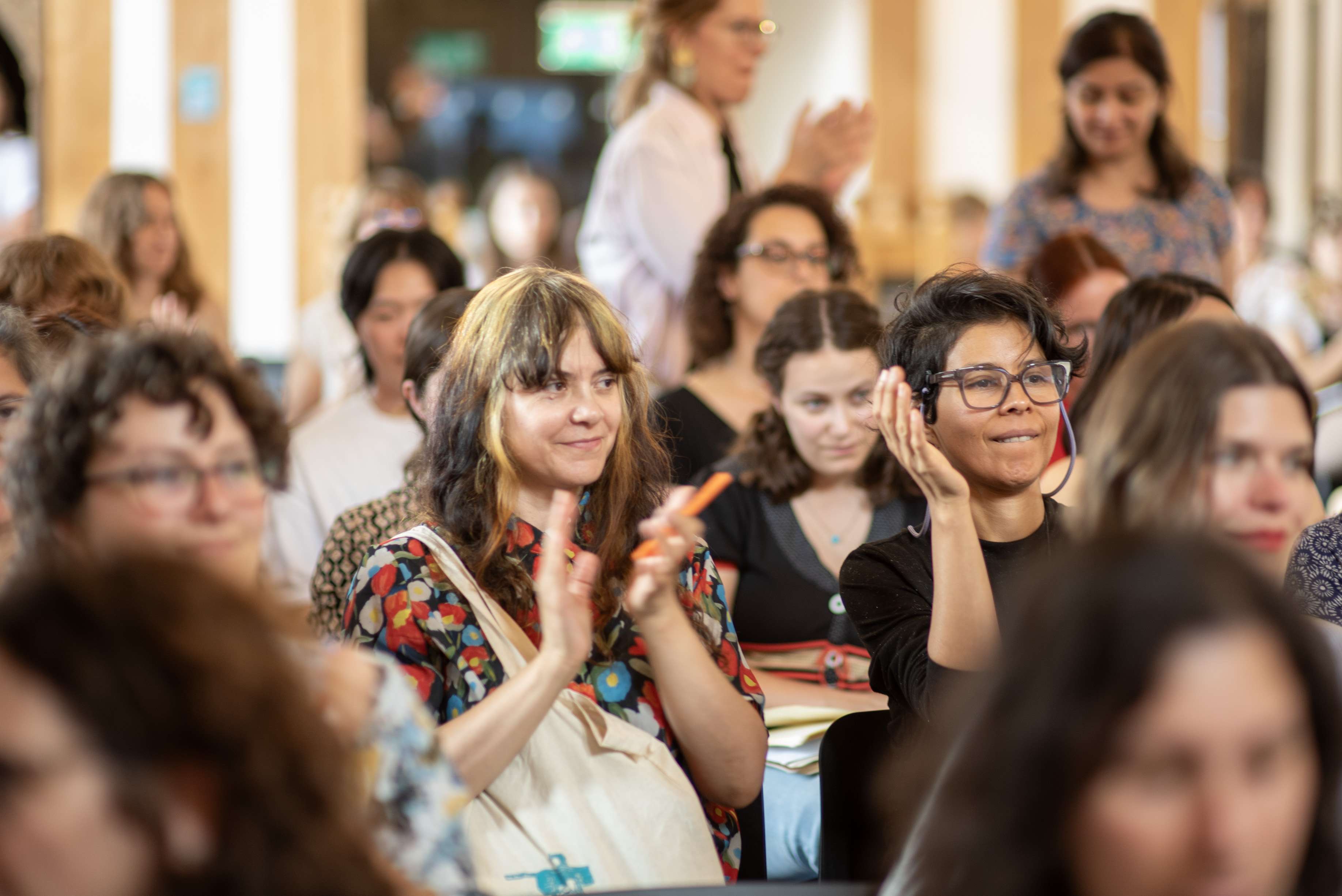 Audience members applaud and smile while seated in a bright room, filled with people. Some are standing in the background.