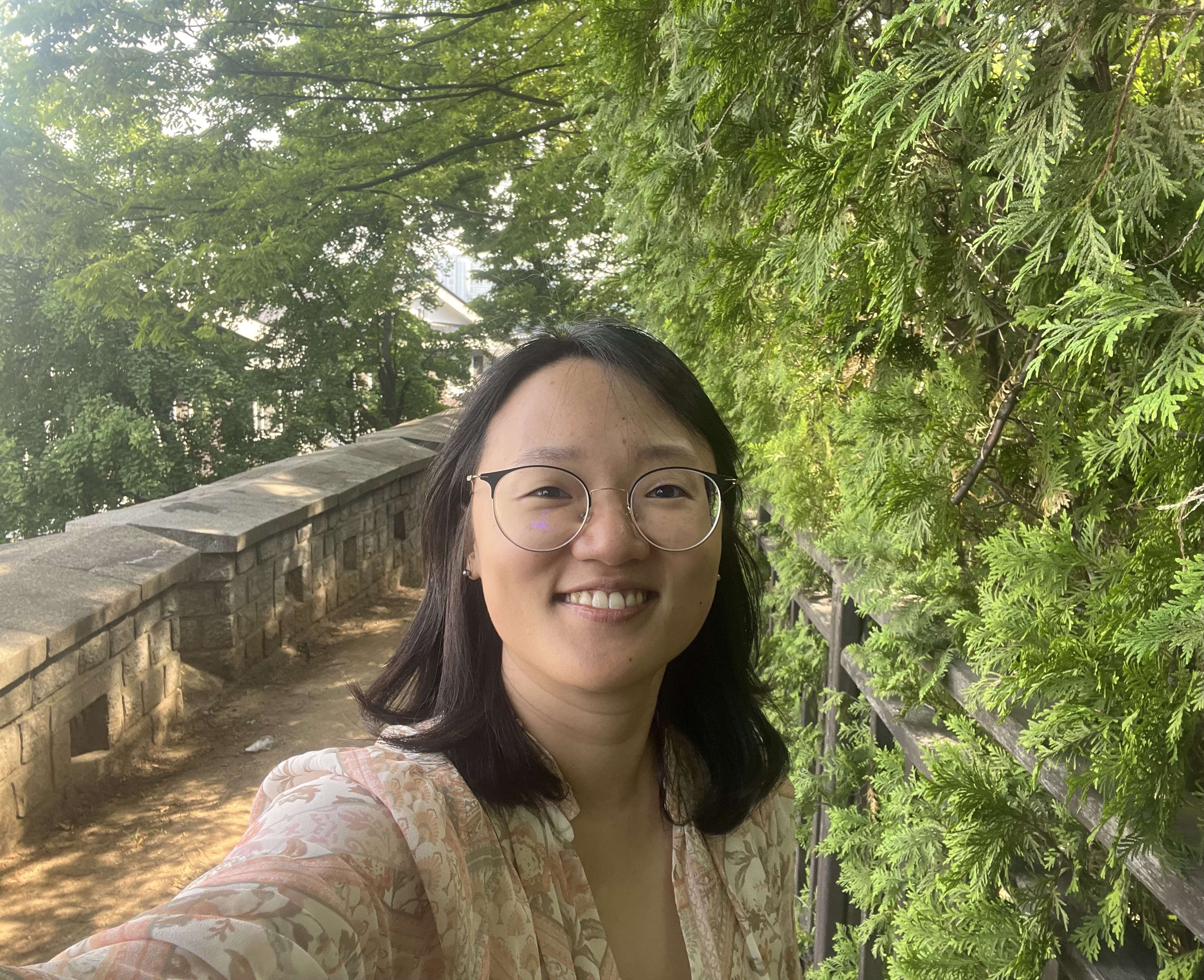 A smiling East Asian woman with glasses and dark hair takes a selfie outdoors next to a stone wall, with lush green trees in the background.