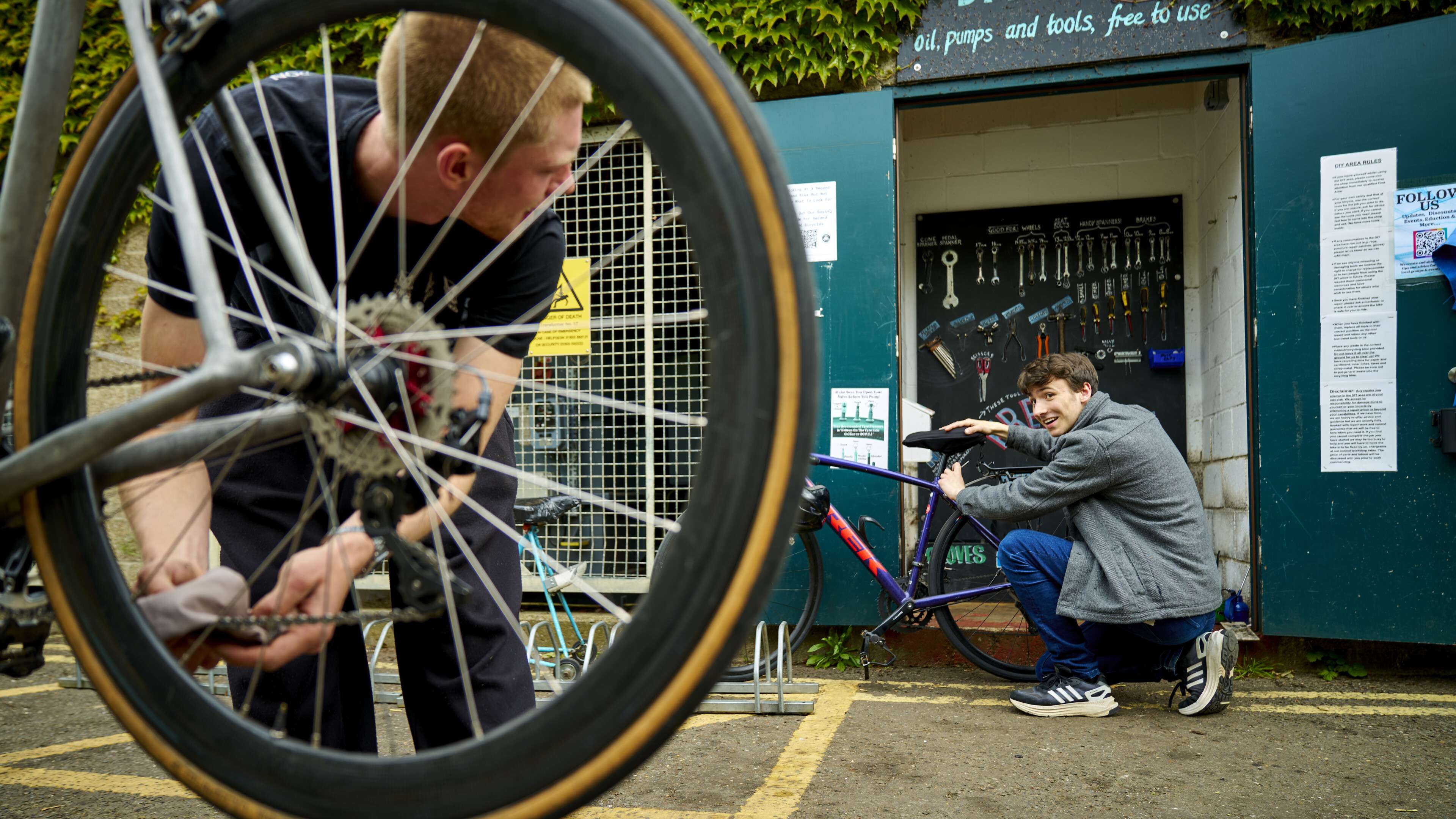 Two men fix their bicycles at a free outdoor repair station, the scene viewed through a bike wheel as one man smiles at the camera.