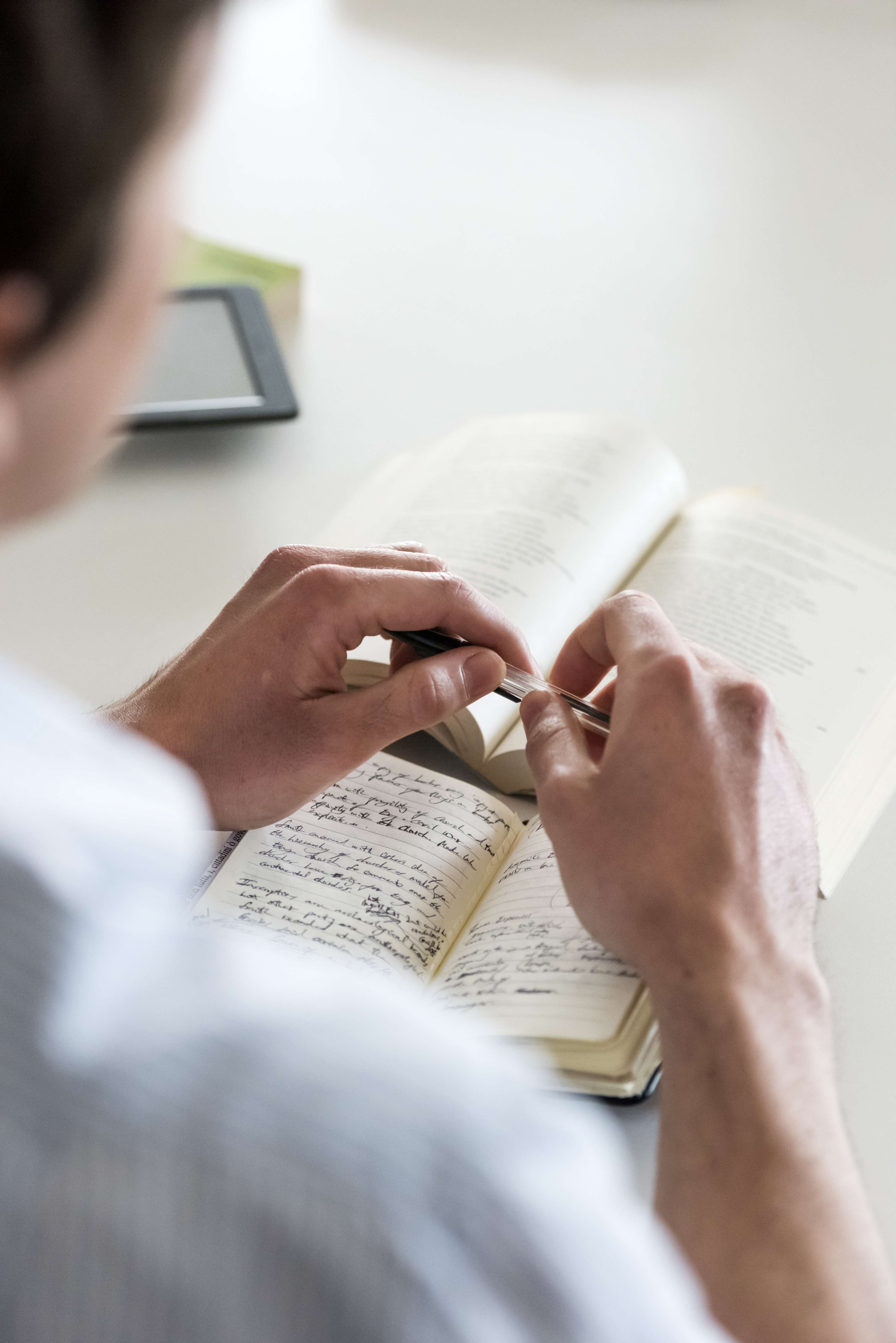 An over-the-shoulder view of a person writing handwritten notes in a notebook while reading a book.