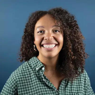 A headshot of a smiling young woman with dark curly hair, wearing a green and white checkered shirt against a solid blue background.