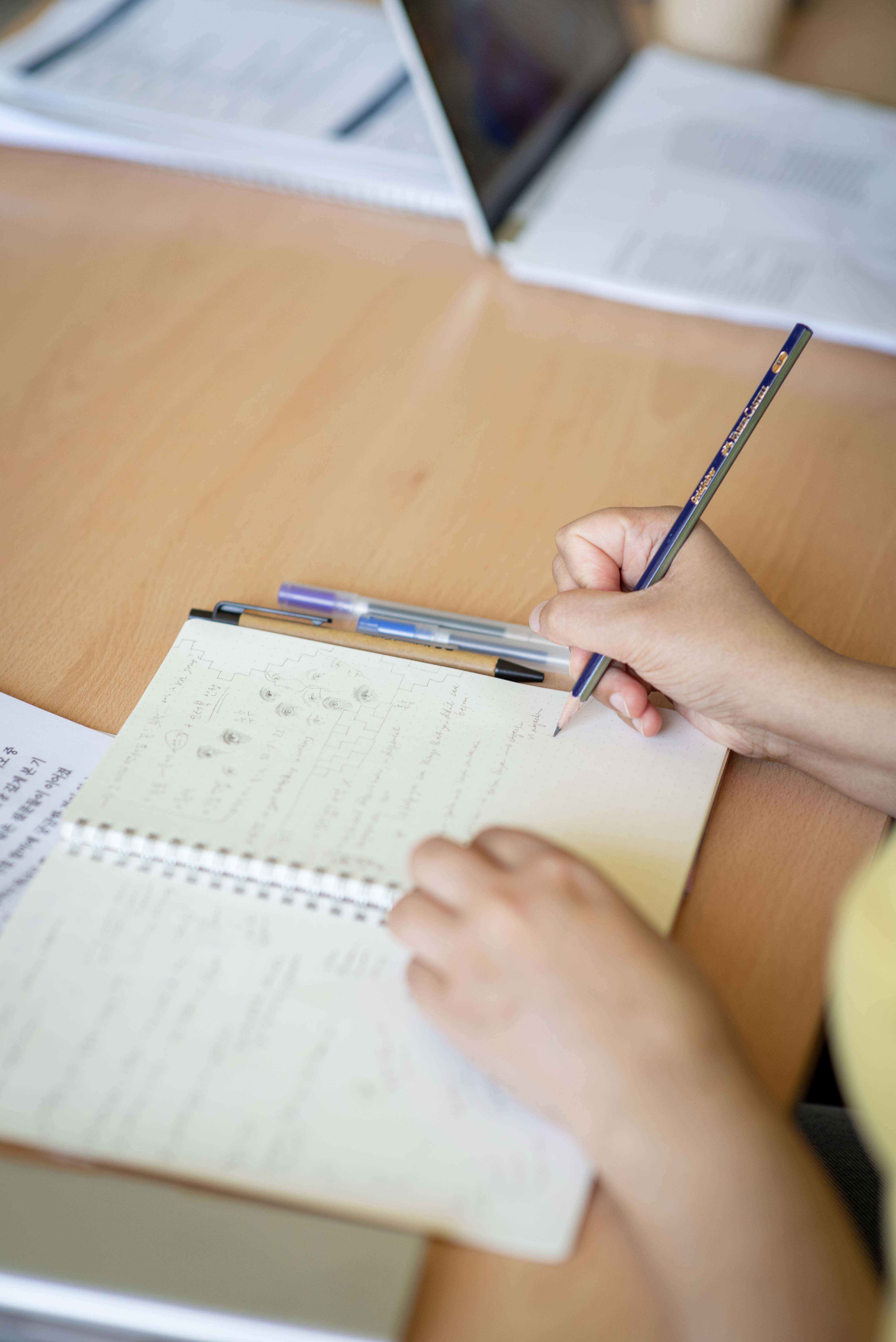Person writing notes in a spiral notebook on a wooden desk, with pens and an open laptop visible in the background.