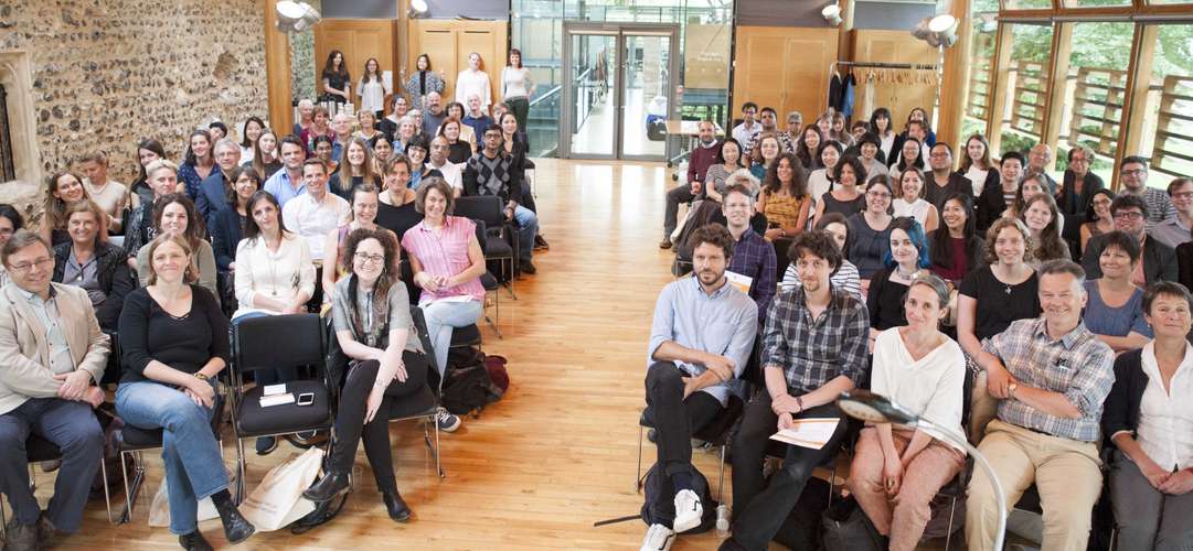 A large group of people seated in a hall, smiling and facing the camera. The room has wooden floors and a brick wall.