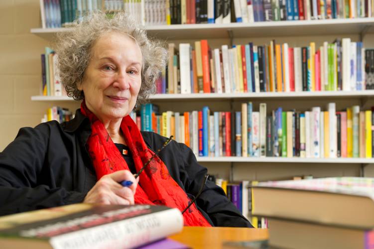 Margaret is an older lady with short grey hair. She had a bright red scarf on and is holding her glasses. She is sitting infront of a book case