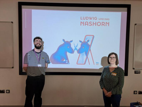 A man and woman stand before a projector screen displaying the title "Ludwig und das Nashorn" and a drawing of a blue rhino looking in a mirror.