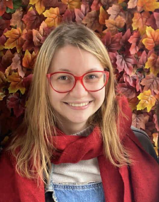 Smiling person with red glasses and scarf, standing in front of a backdrop of colorful autumn leaves.