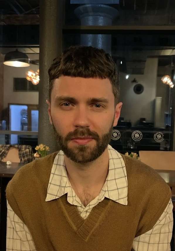 A man with short brown hair, a fringe, and a beard looks directly at the camera. He wears a brown sweater vest over a checkered collared shirt.